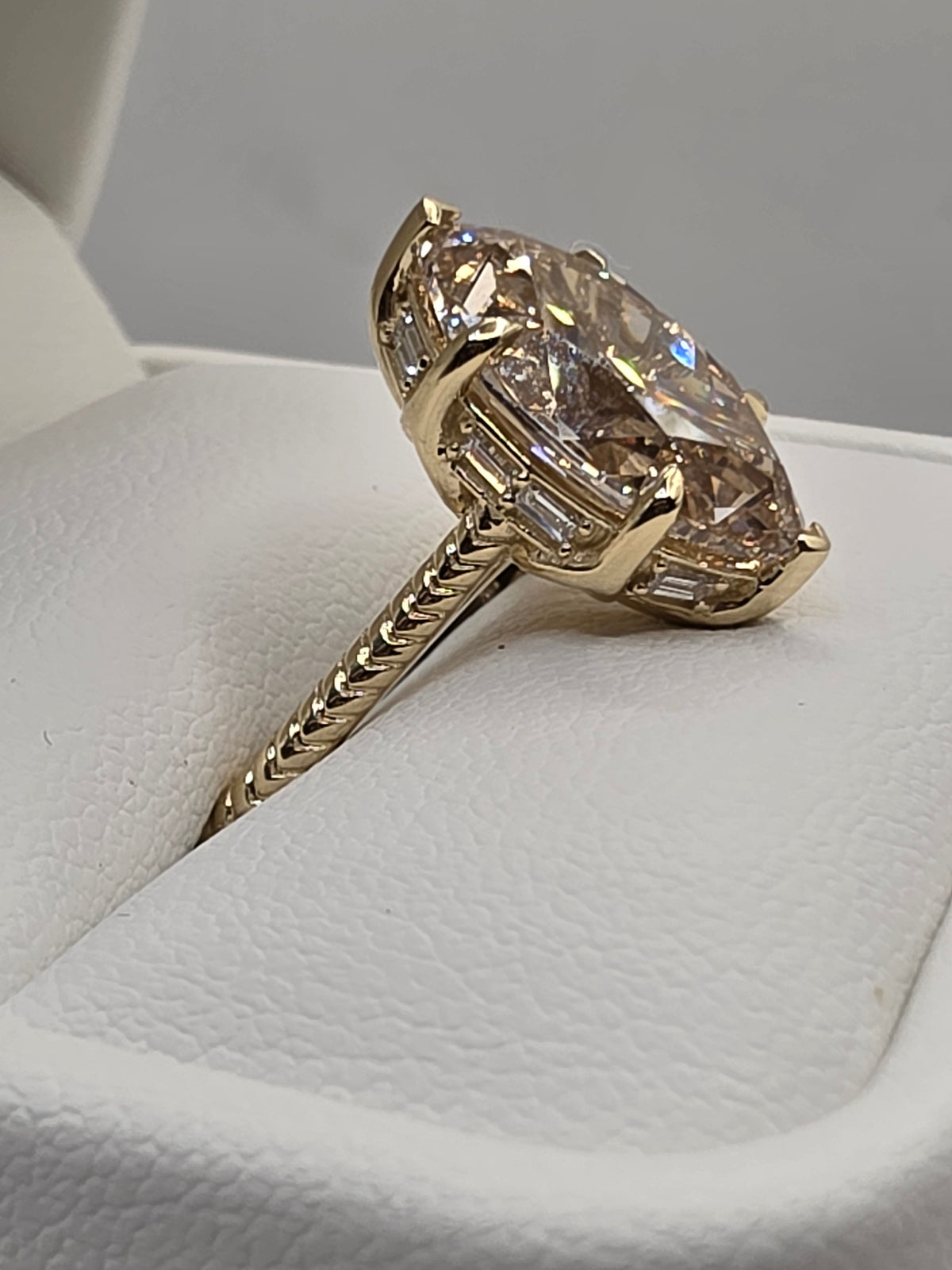 A yellow gold ring with a large marquise-cut clear gemstone and smaller side stones, displayed on a white textured ring box.