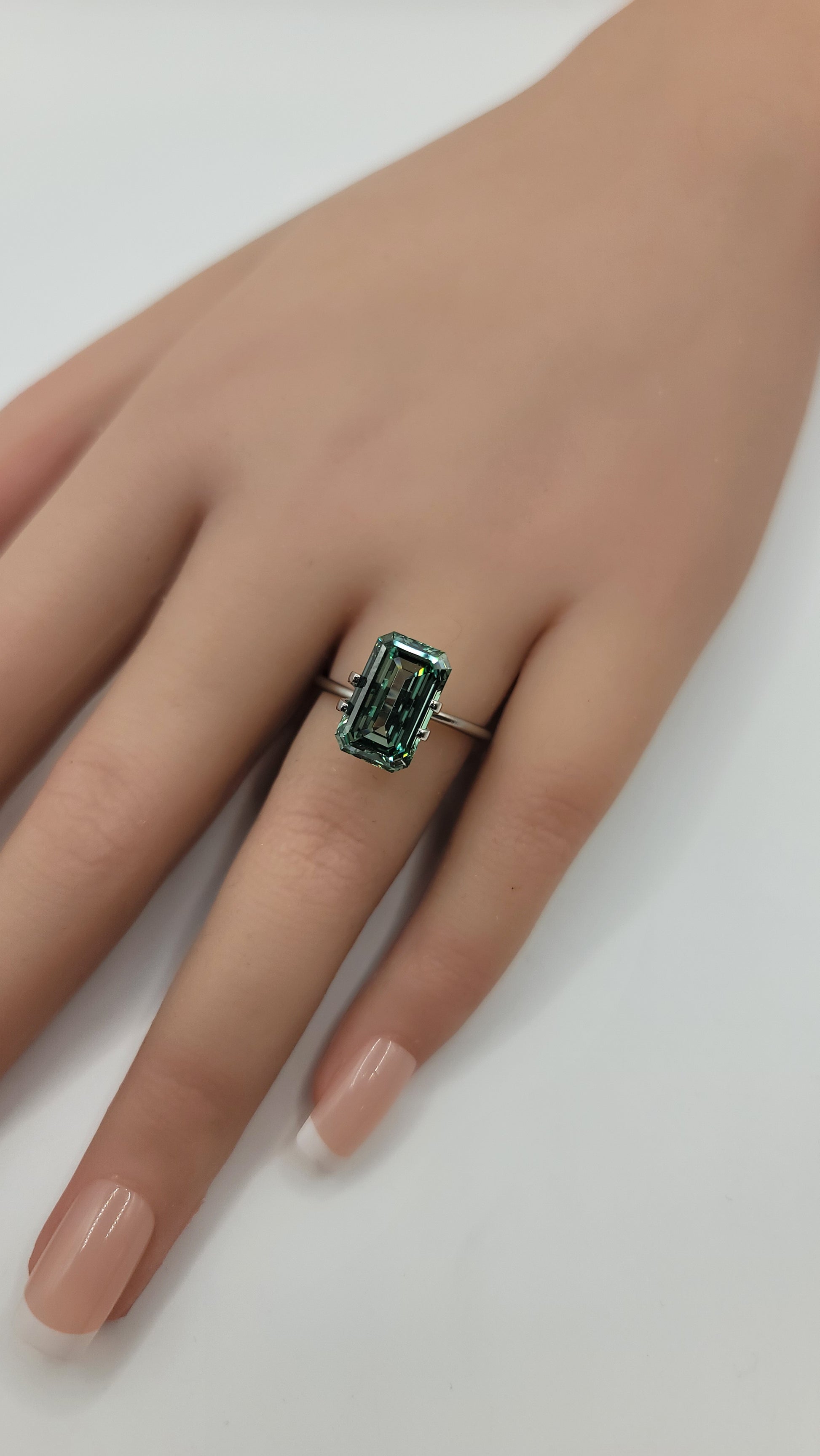 A close-up of a hand wearing a silver ring with a rectangular emerald-cut green diamond, set against a plain white background.