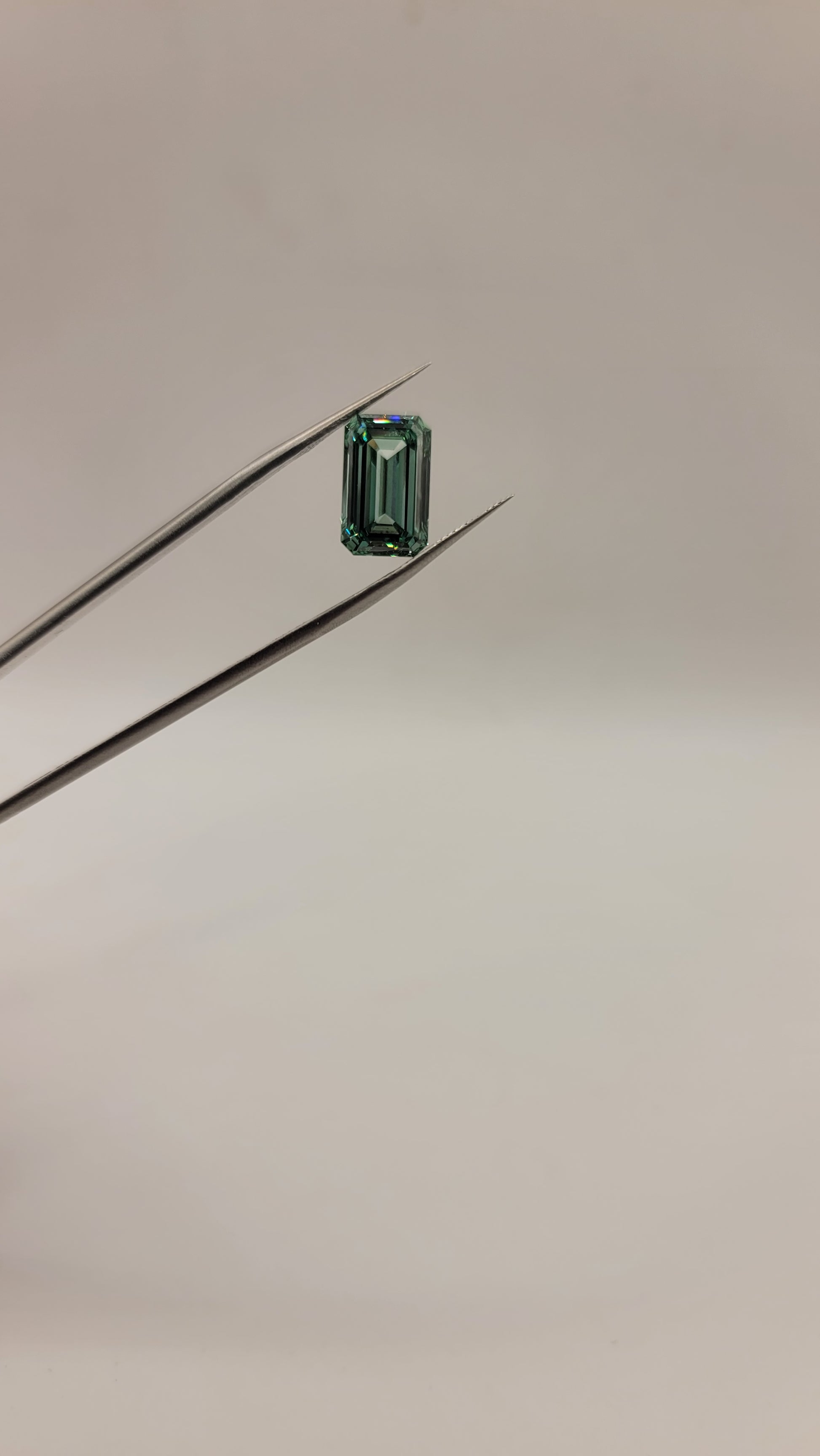 A small rectangular emerald-cut green diamond held between two metal tweezers against a plain light background.
