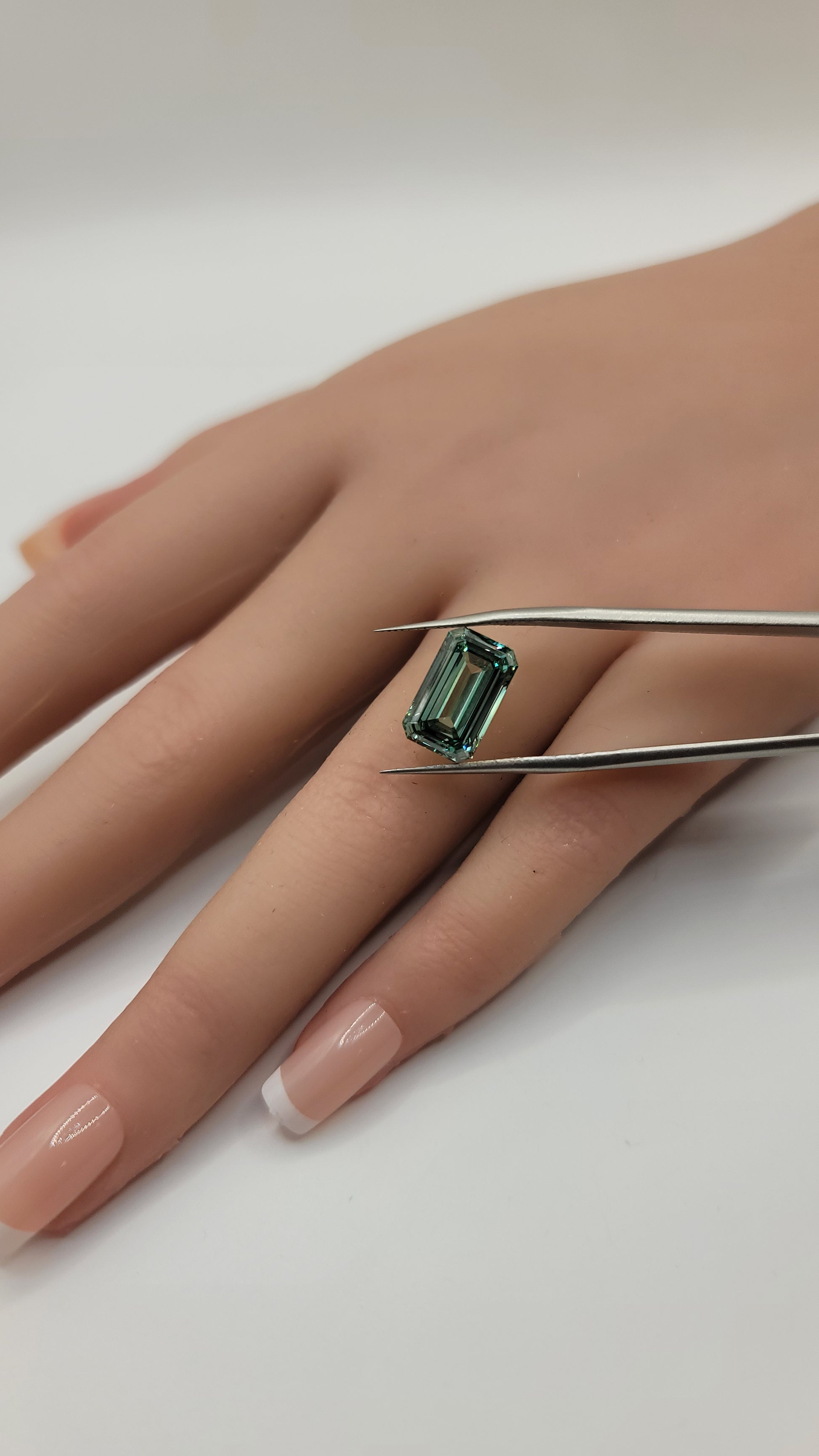 A close-up of a hand holding a small rectangular green diamond with metal tweezers against a plain light background.