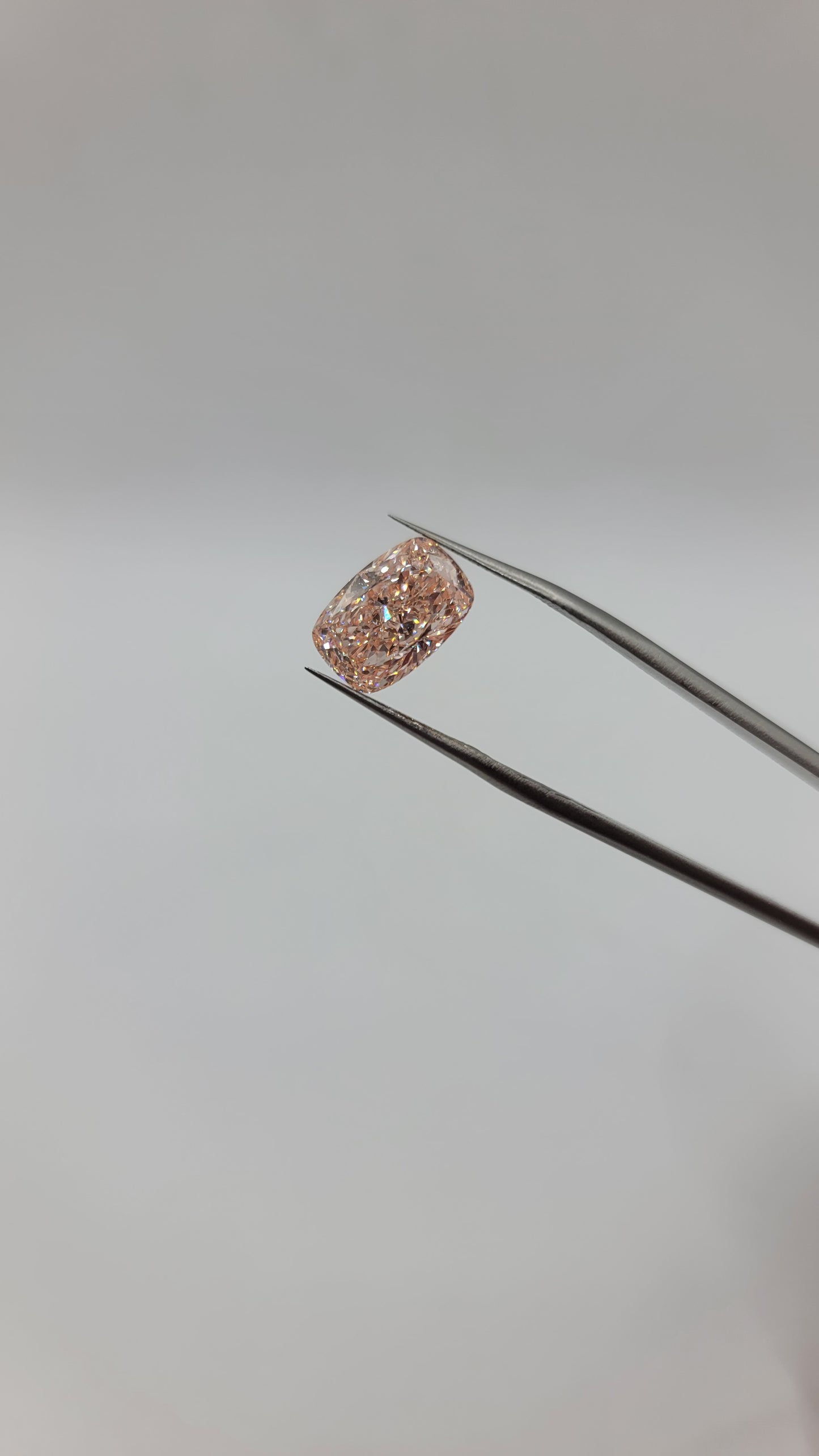Lab grown pink diamond held in the tips of metal tweezers against a plain light gray background.