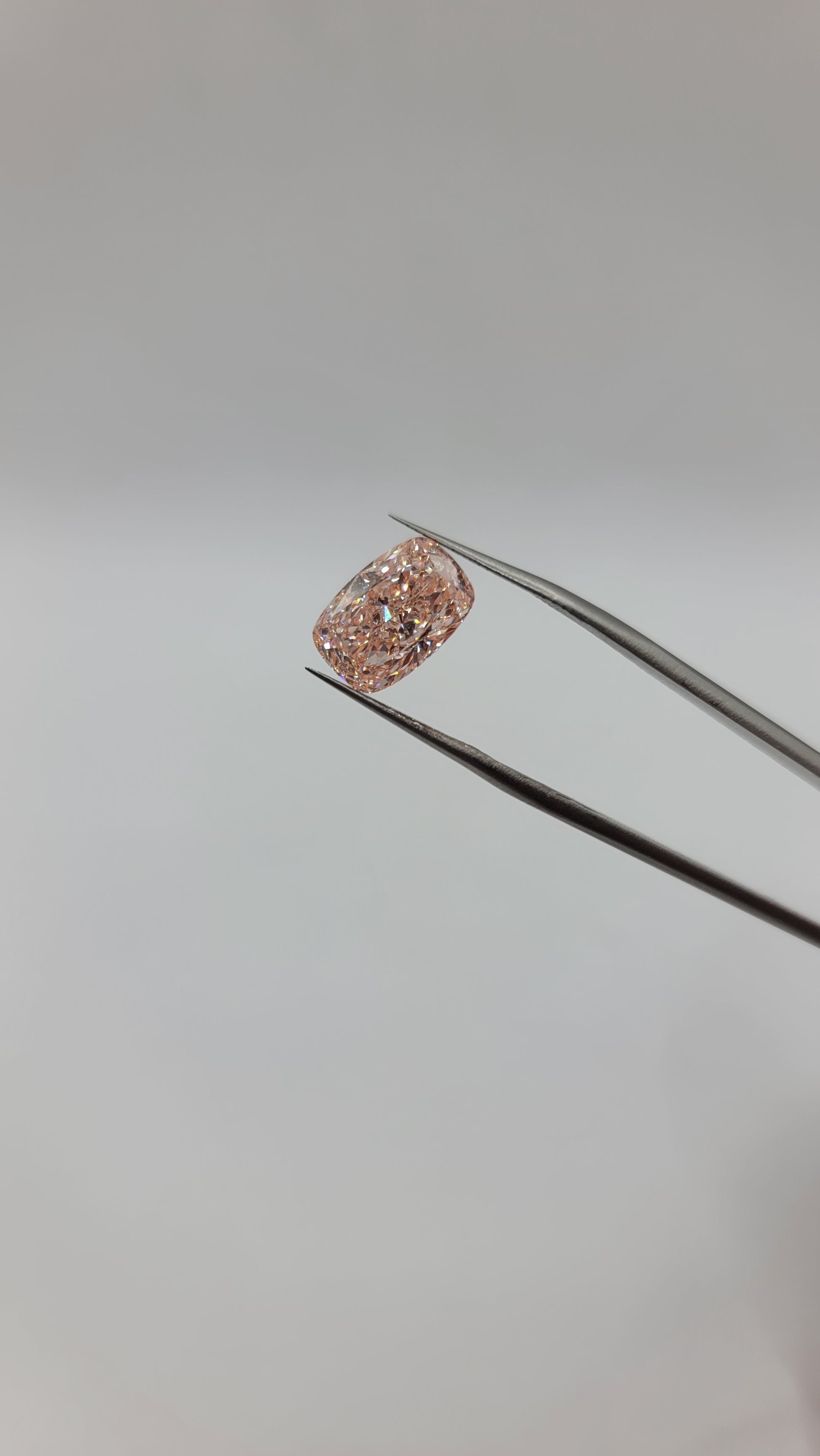 Lab grown pink diamond held in the tips of metal tweezers against a plain light gray background.