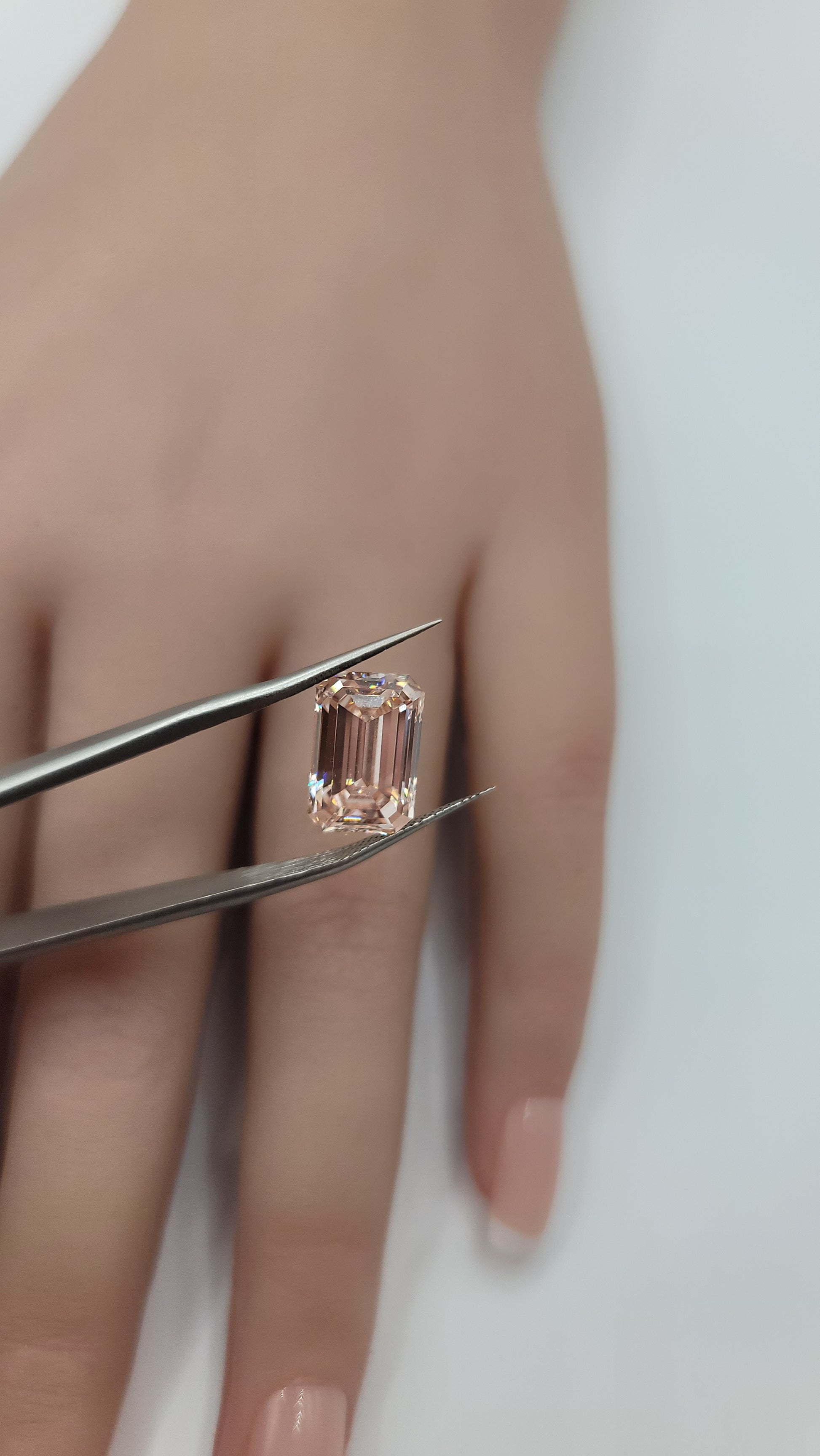 A pink emerald-cut lab diamond held between metal tweezers over a person's fingers.
