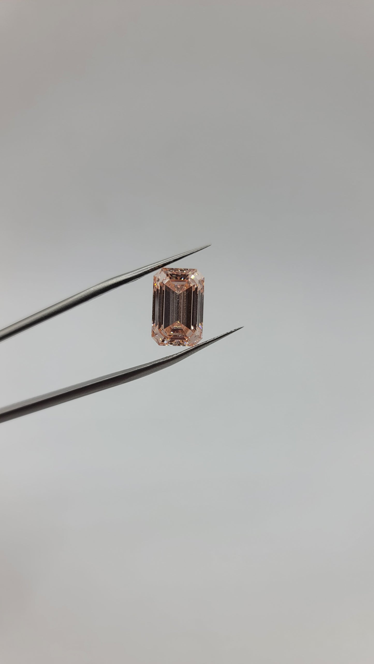 A rectangular emerald-cut pink lab diamond held between two metal tweezers against a plain light gray background.