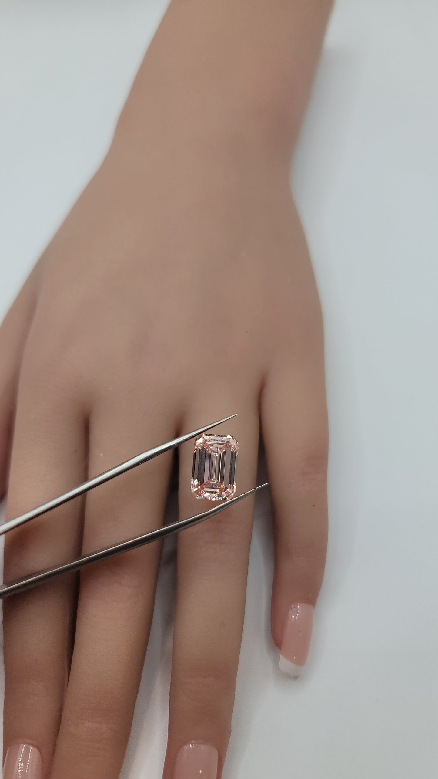 A close-up of a hand holding a rectangular emerald-cut lab diamond between metal tweezers against a plain light background.