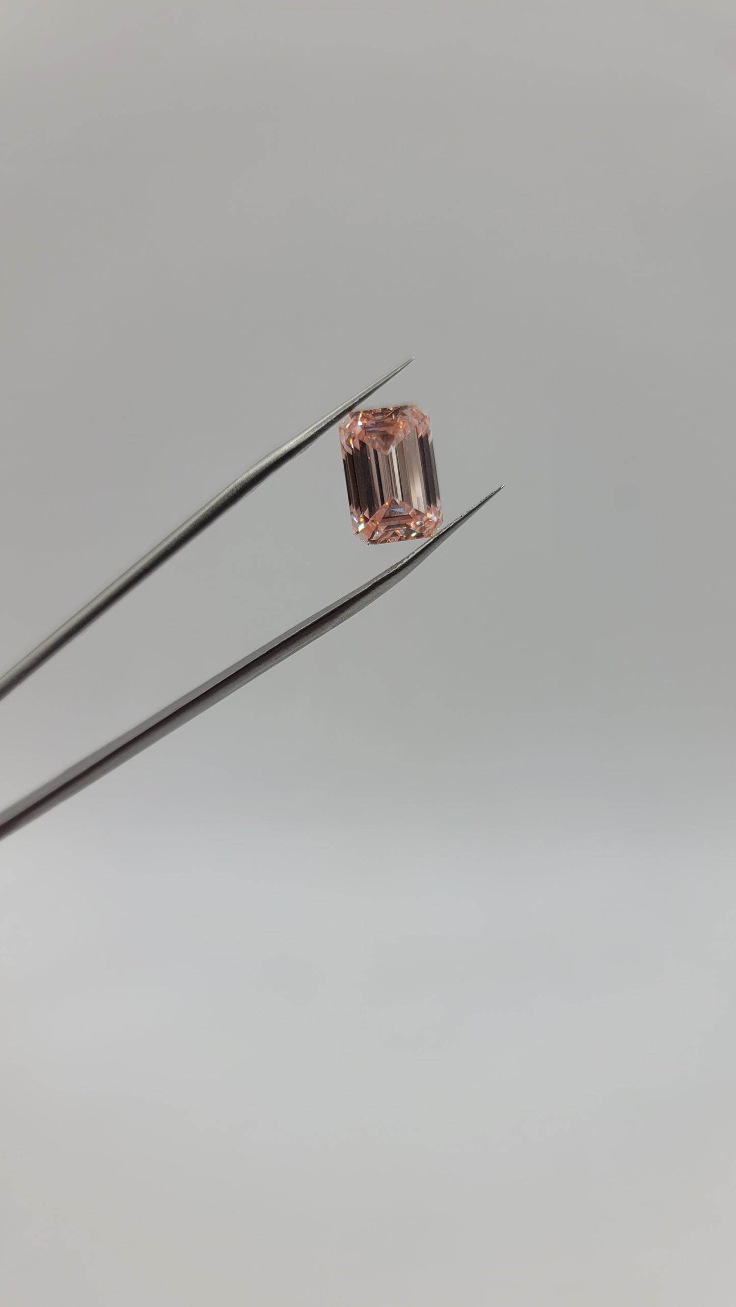 A small rectangular pink lab diamond held between metal tweezers against a plain light gray background.