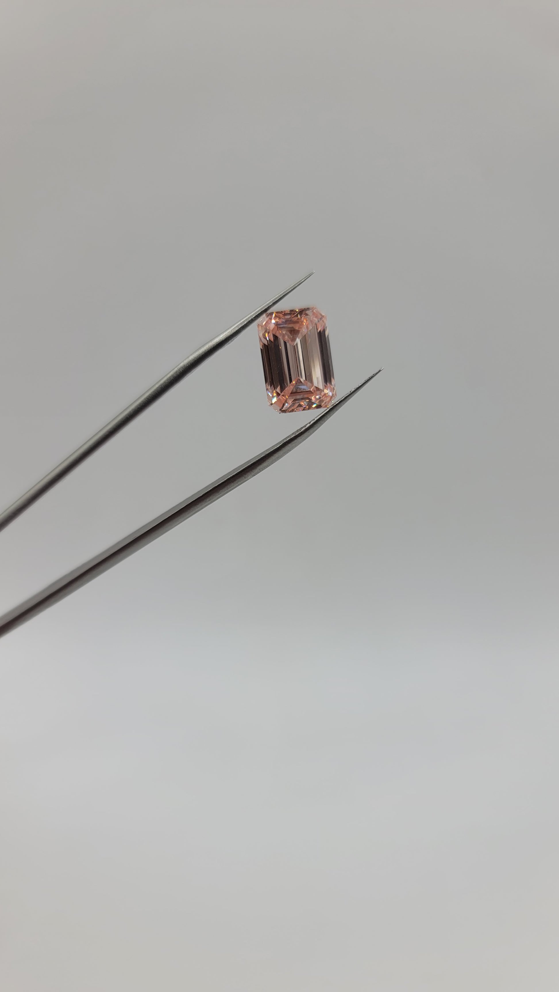 A small rectangular pink lab diamond held between metal tweezers against a plain light gray background.