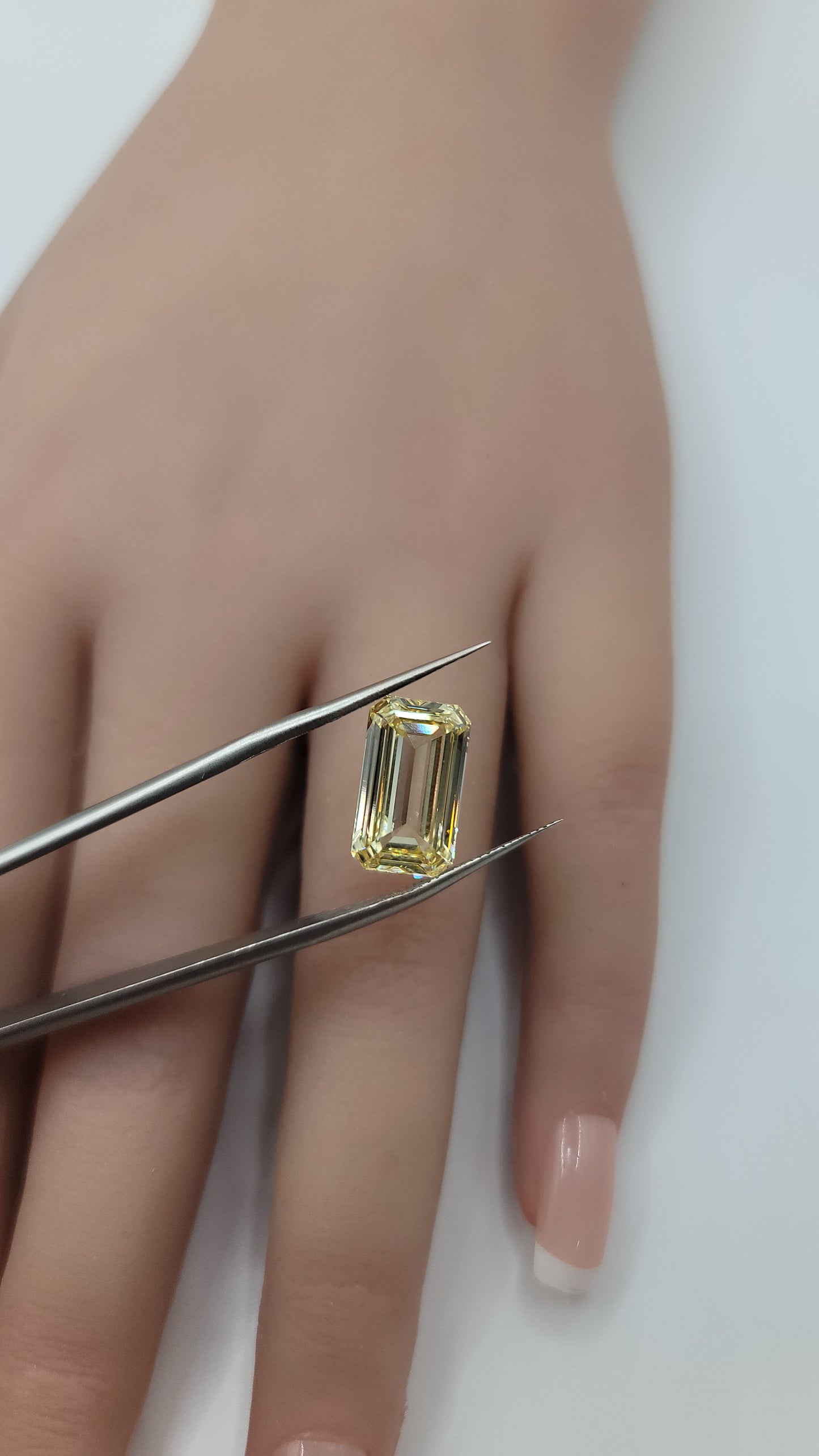A yellow emerald-cut lab diamond held by metal tweezers against a person's hand, showing the clear stone with its yellow color.