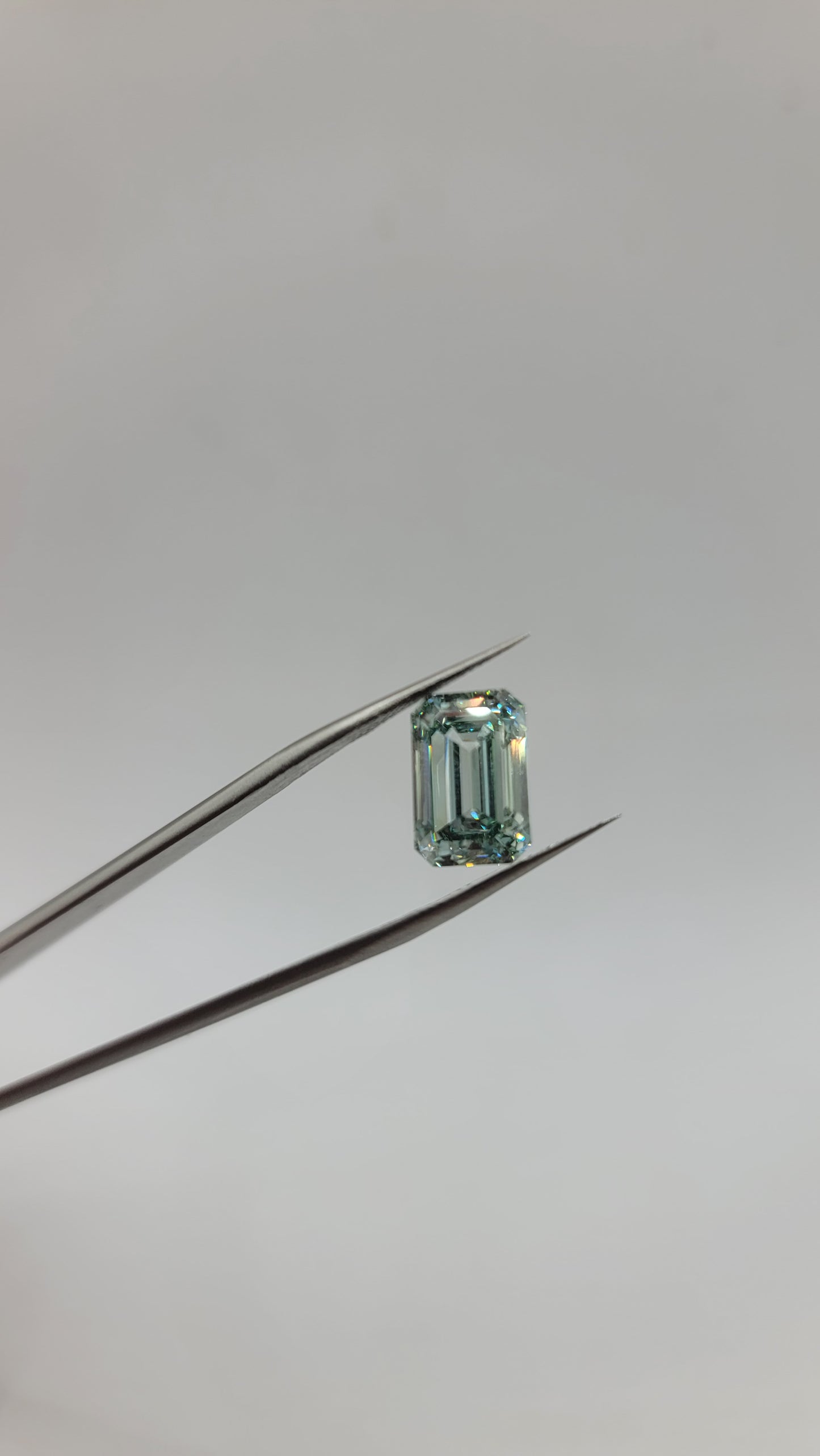 A rectangular emerald-cut green lab diamond held between metal tweezers against a plain light gray background.