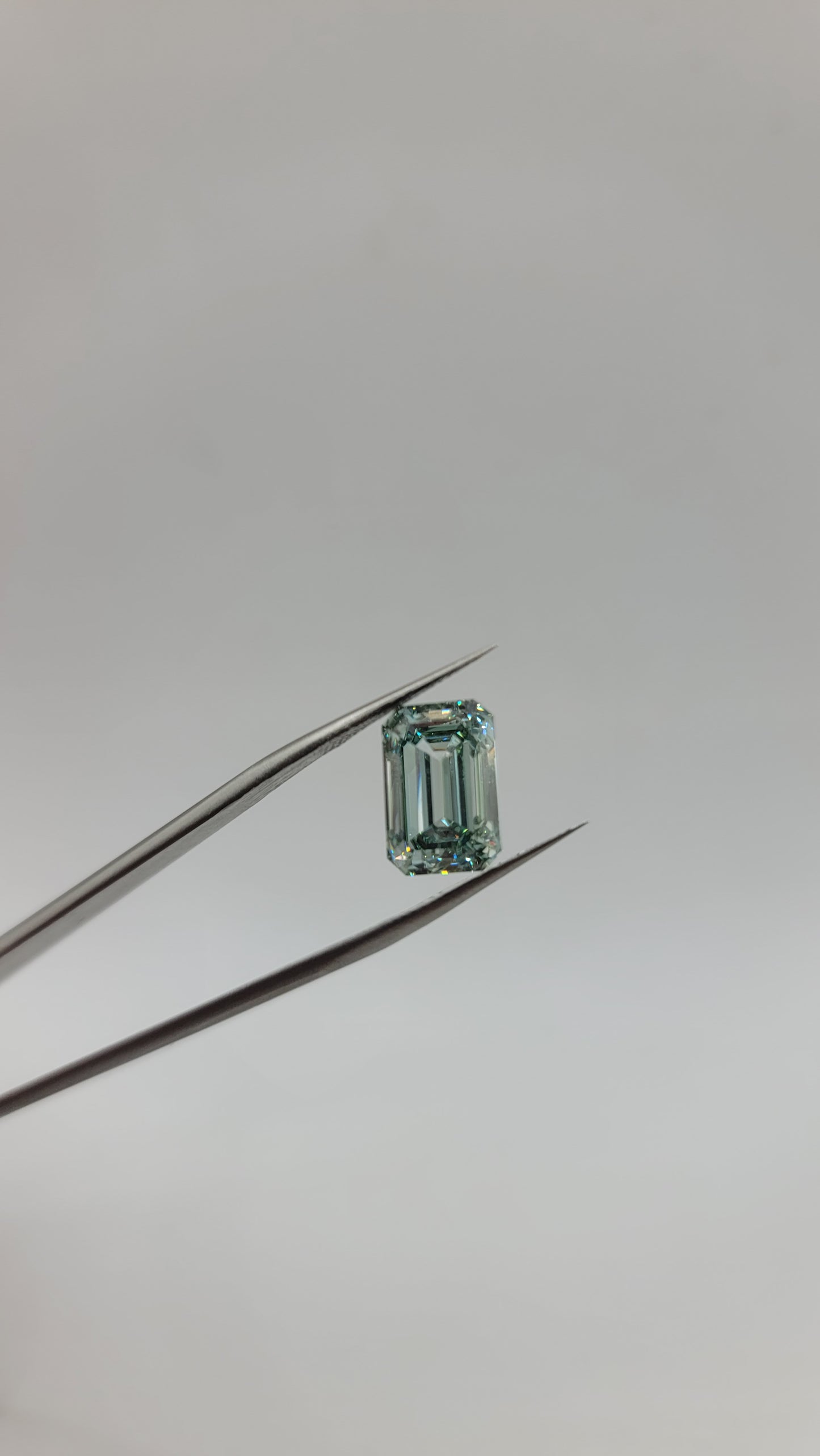 A small rectangular emerald-cut green lab diamond held between metal tweezers against a plain light gray background.