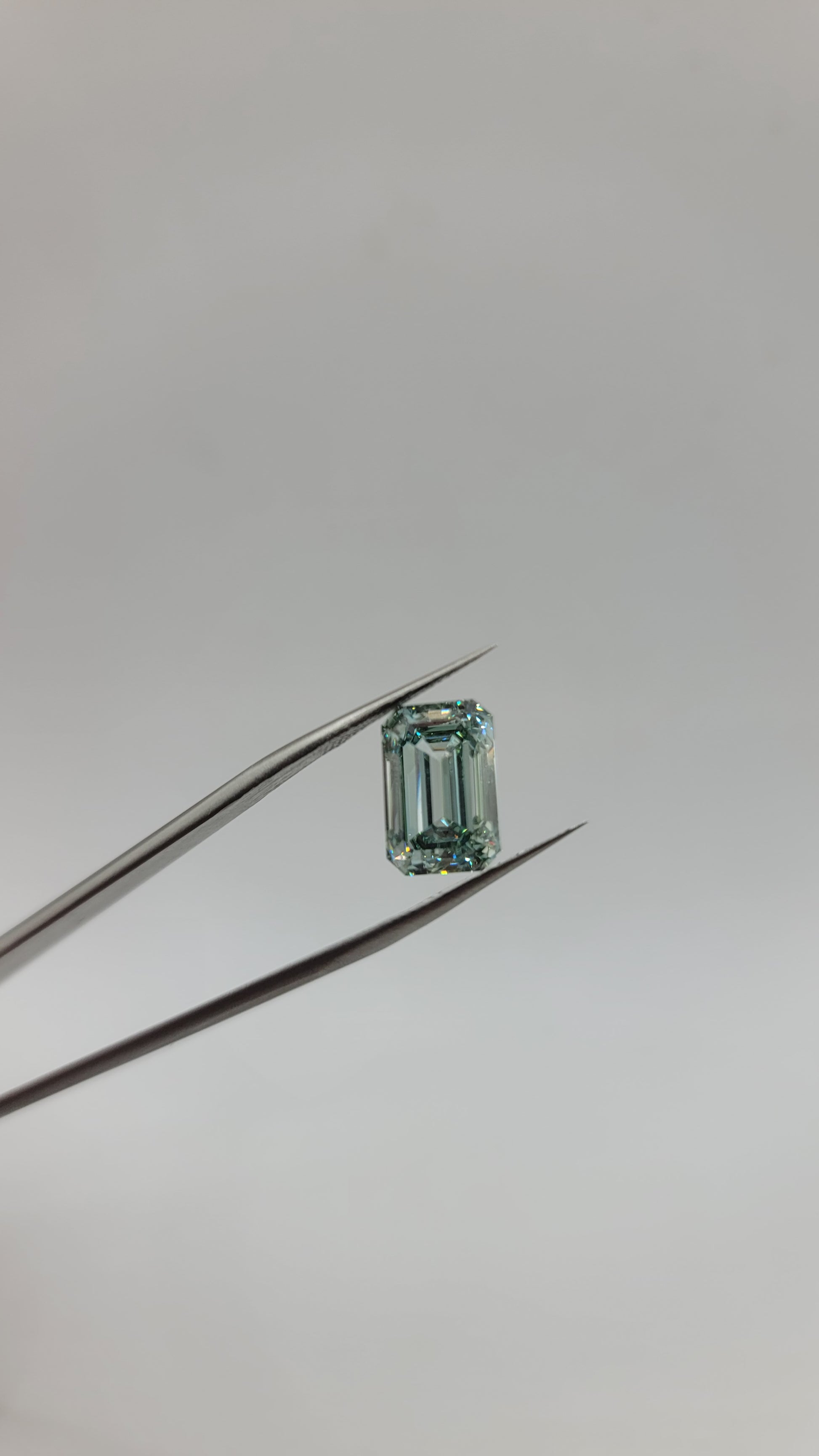 A small rectangular emerald-cut green lab diamond held between metal tweezers against a plain light gray background.