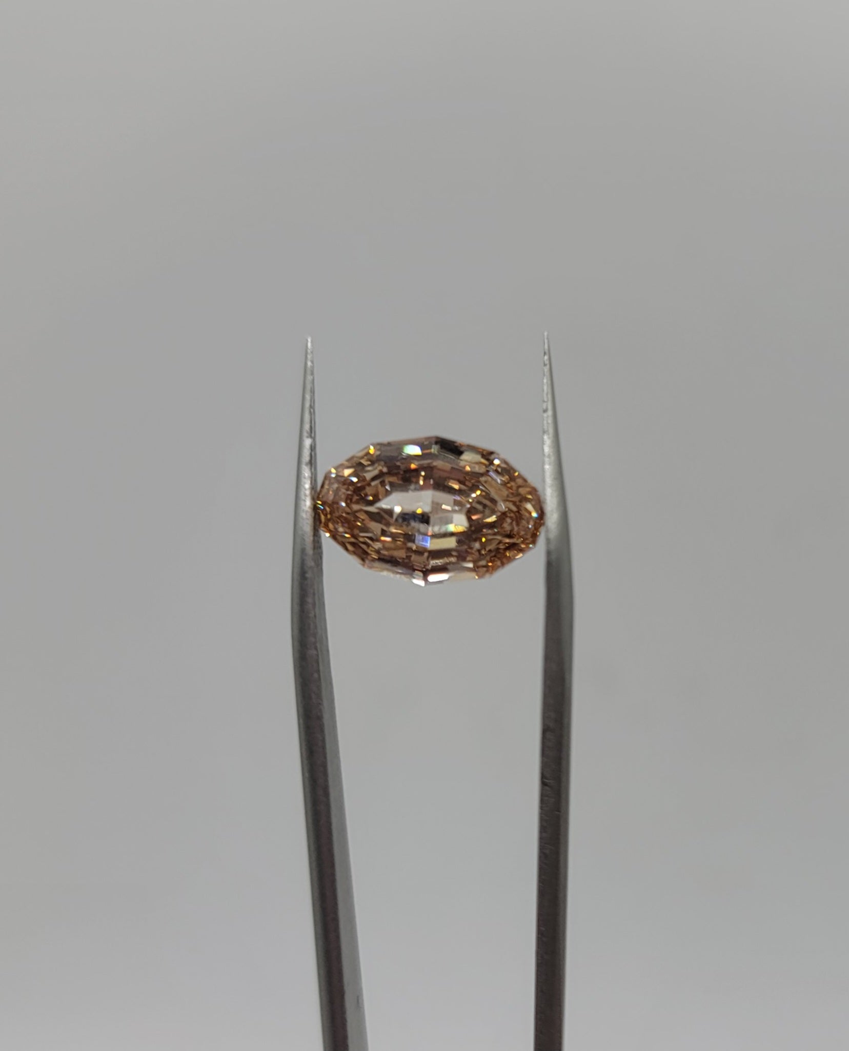 A oval lab diamond held in the center of silver tweezers against a solid gray background.