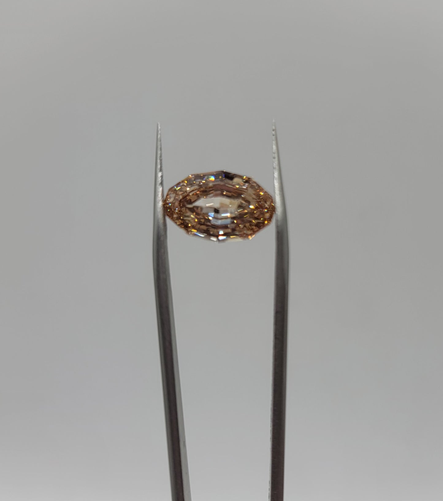 A oval lab diamond held in the center of silver tweezers against a neutral gray background.
