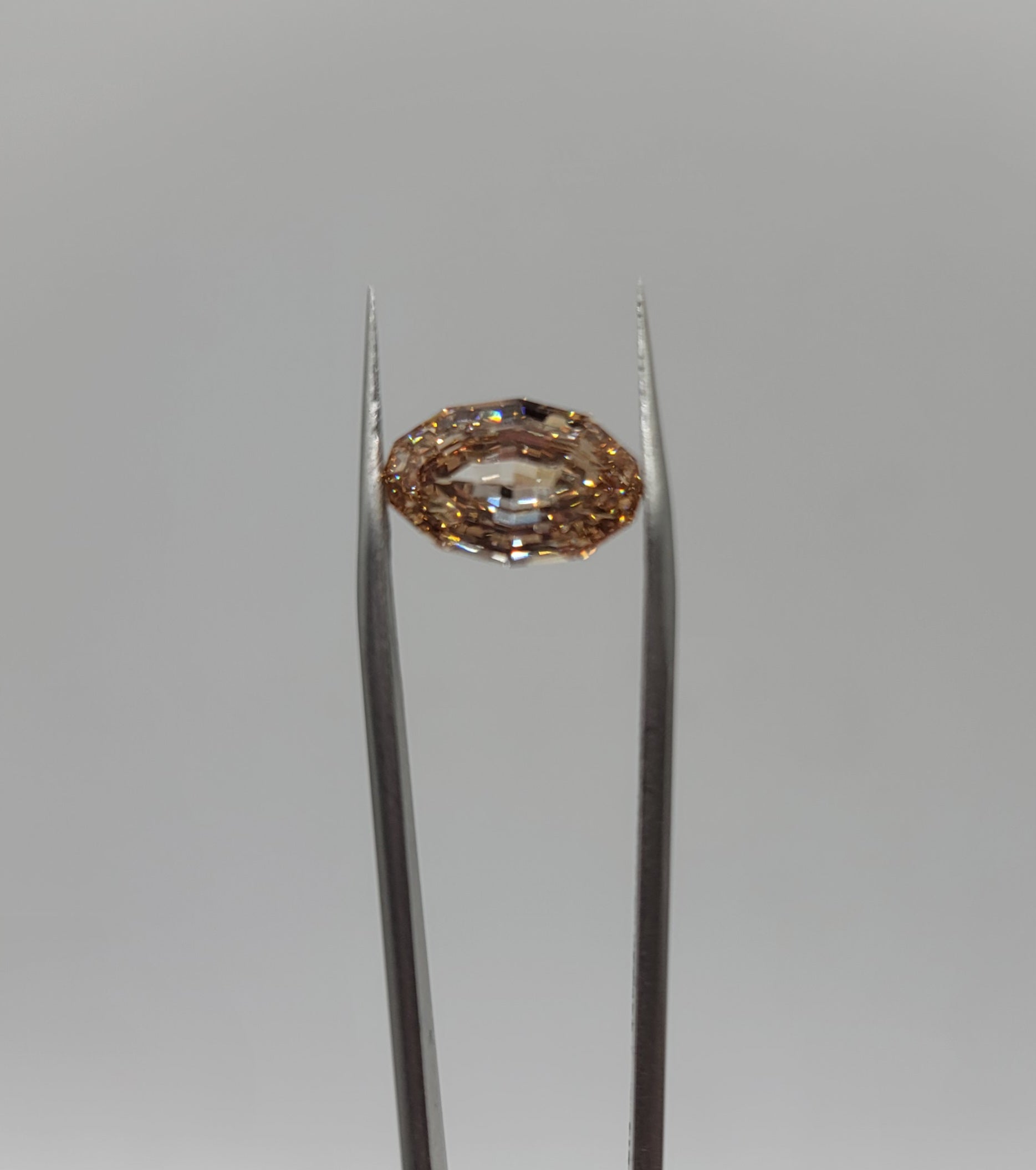 A oval lab diamond held in the center of silver tweezers against a neutral gray background.