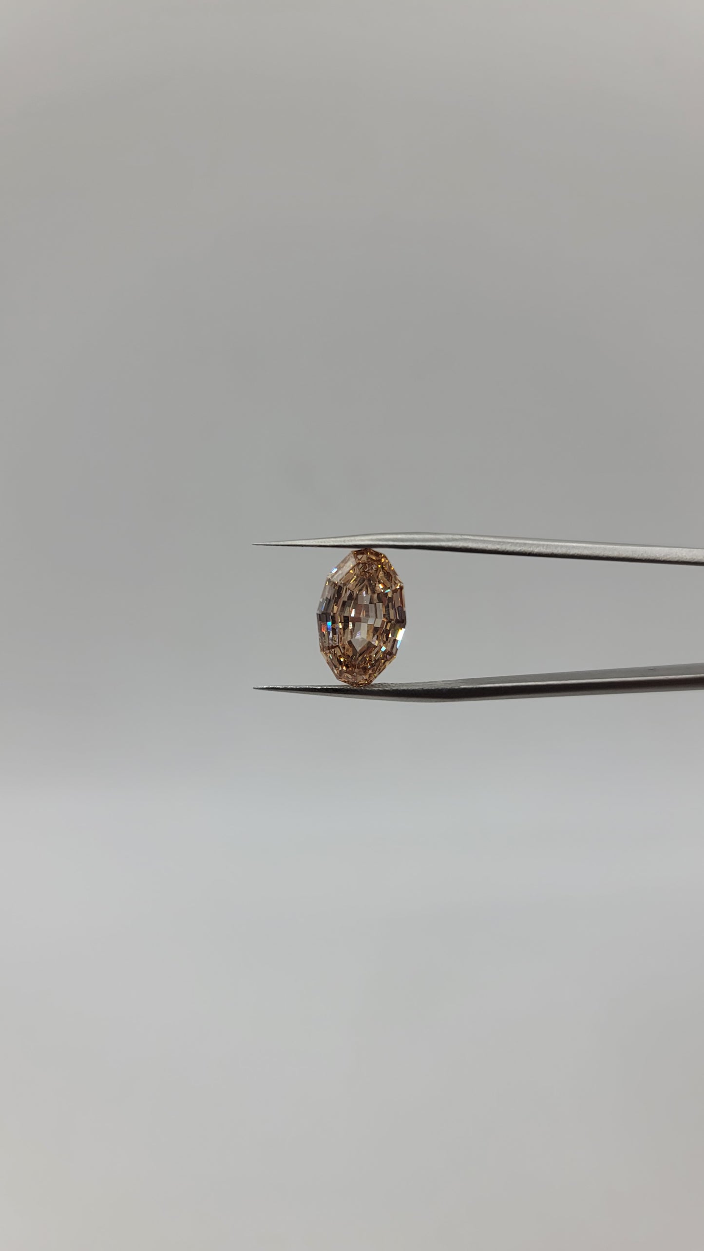 A oval lab diamond between two metal tweezers against a plain light gray background.