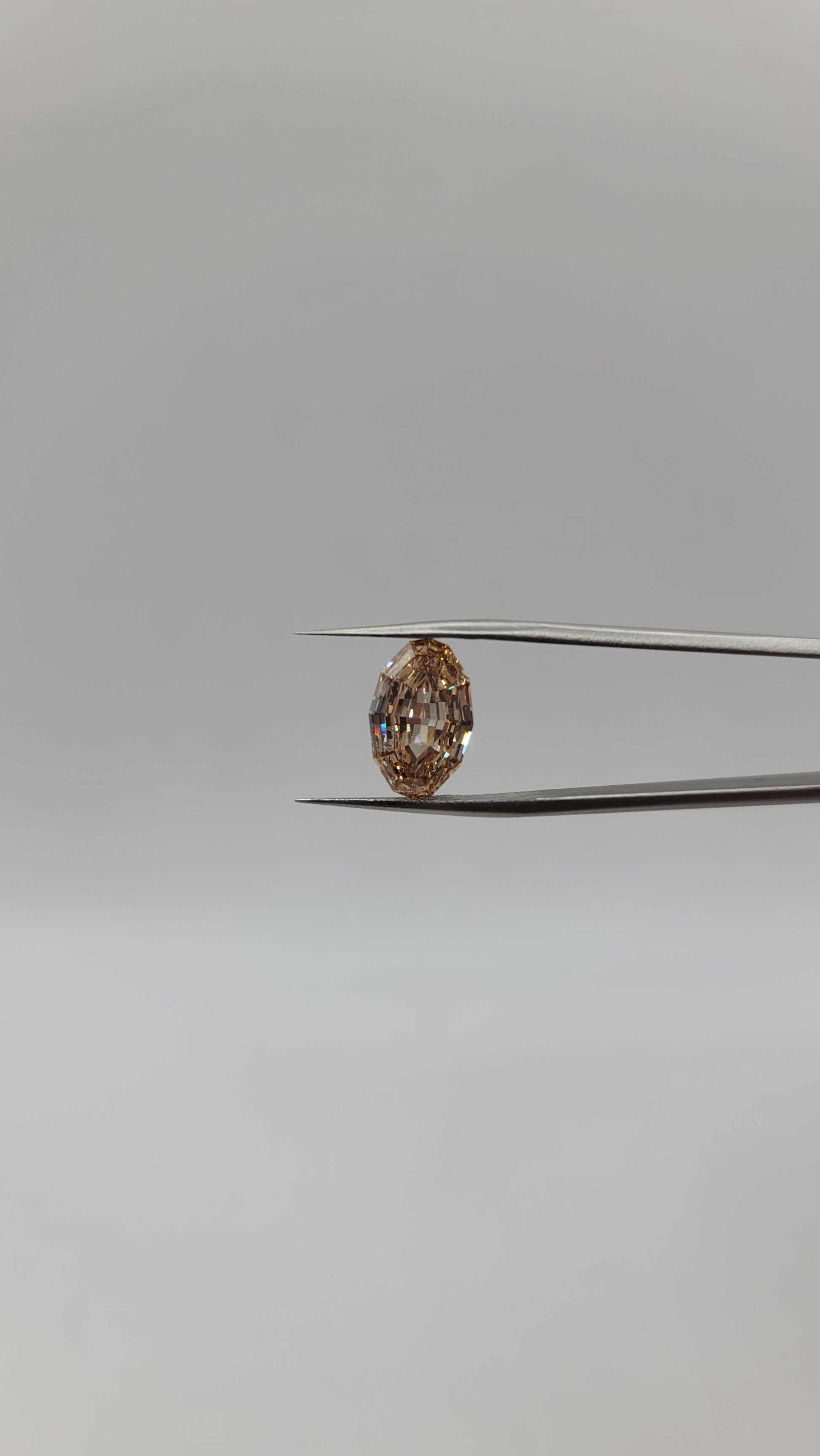A oval lab diamond between two metal tweezers against a plain light gray background.