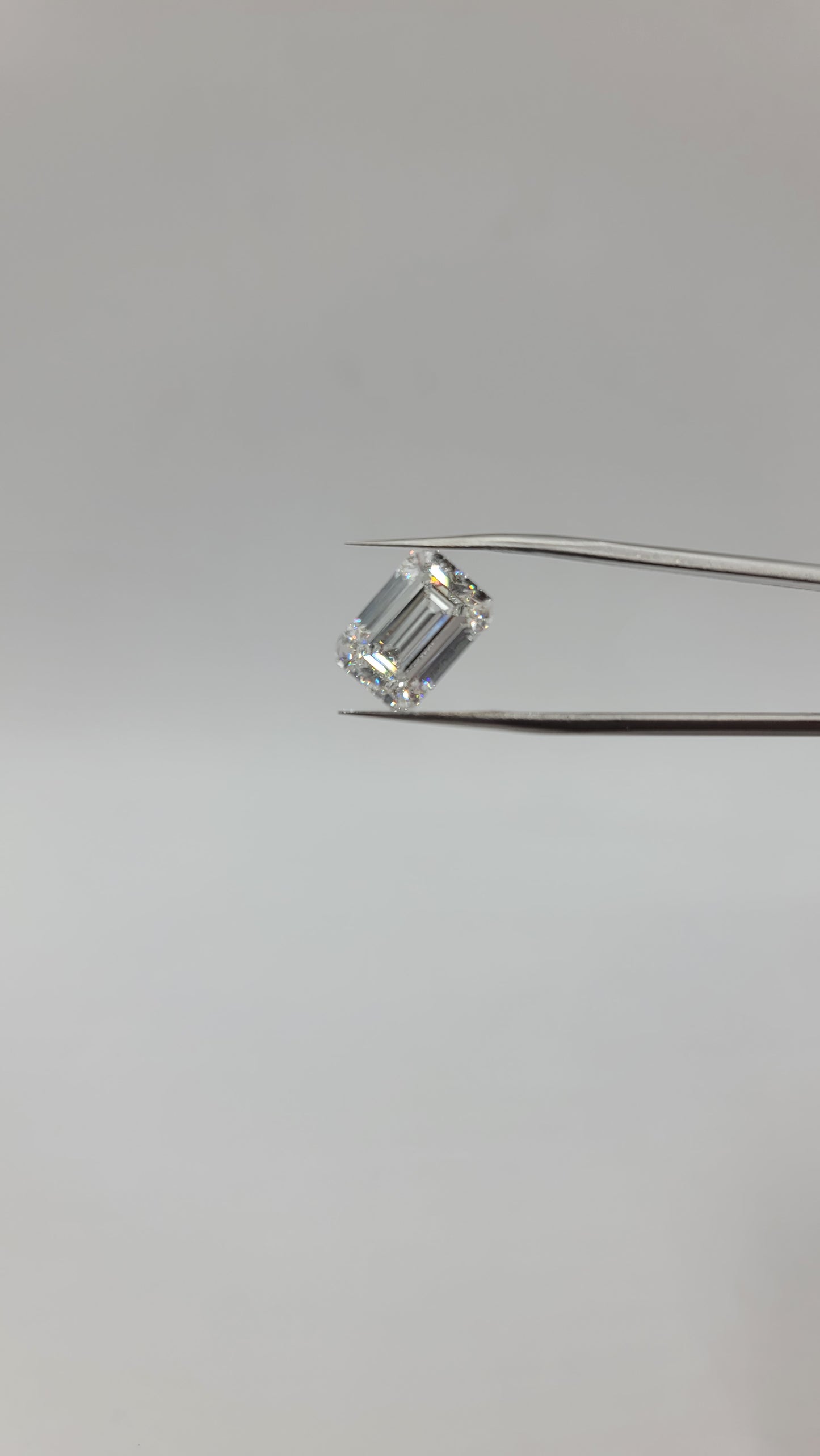 A emerald cut lab diamond held between two metal tweezers against a plain light gray background.