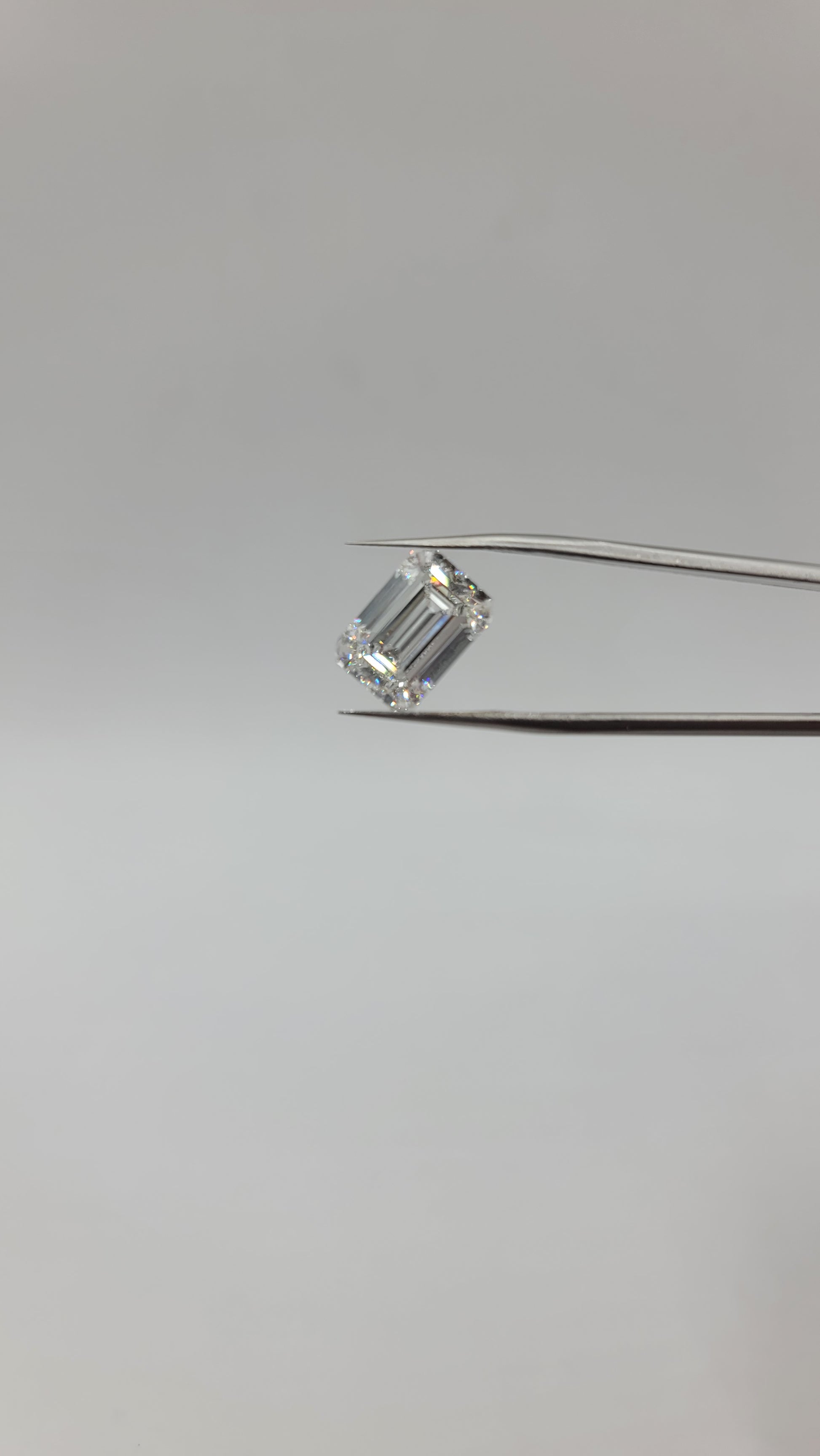 A emerald cut lab diamond held between two metal tweezers against a plain light gray background.