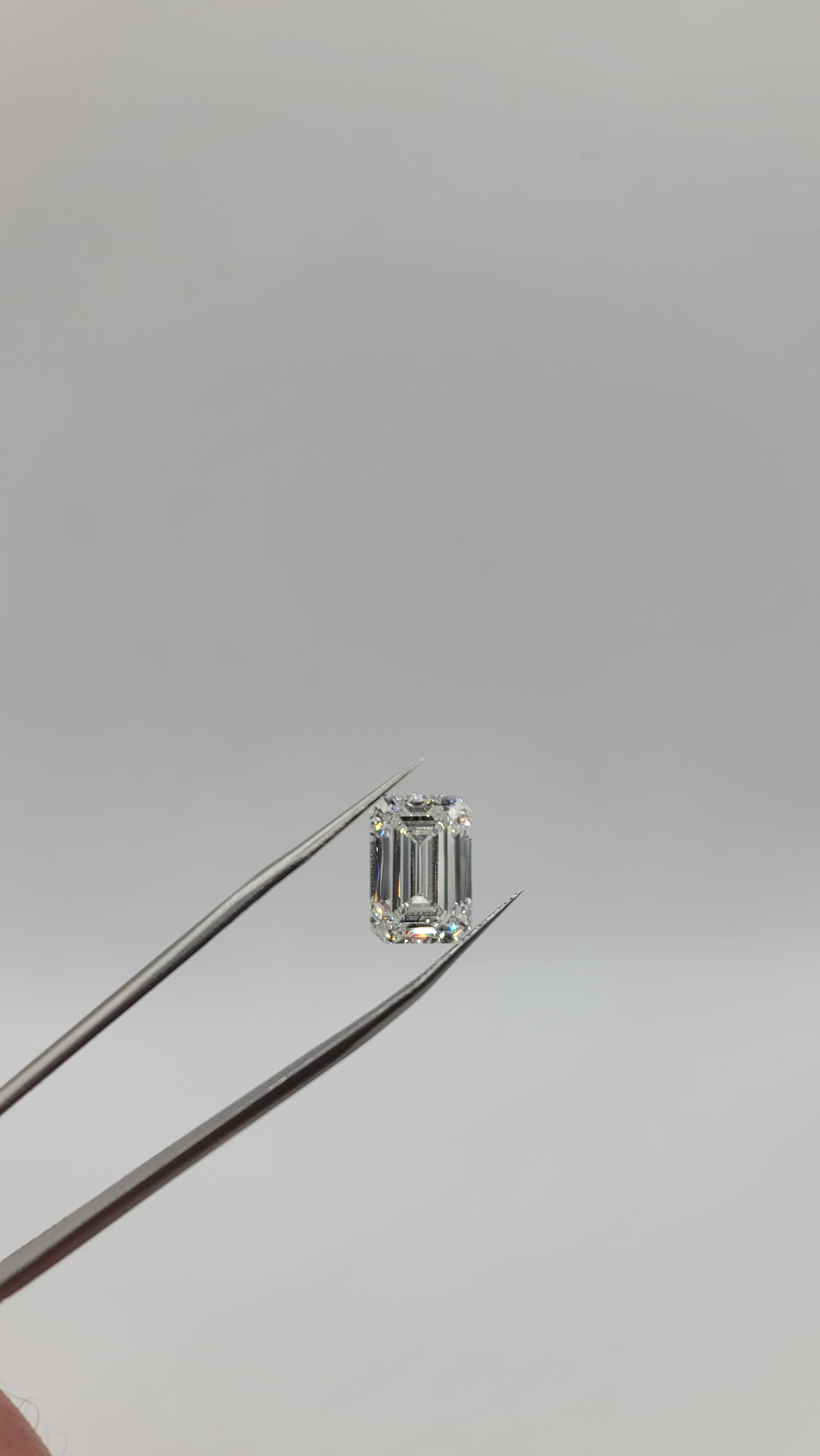 An emerald-cut lab diamond held by metal tweezers against a plain light gray background.