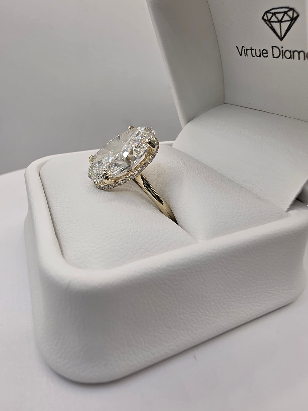 A yellow gold ring with a large oval clear gemstone and a halo of smaller stones, displayed inside an open white jewelry box branded 'Virtue Diamonds'.
