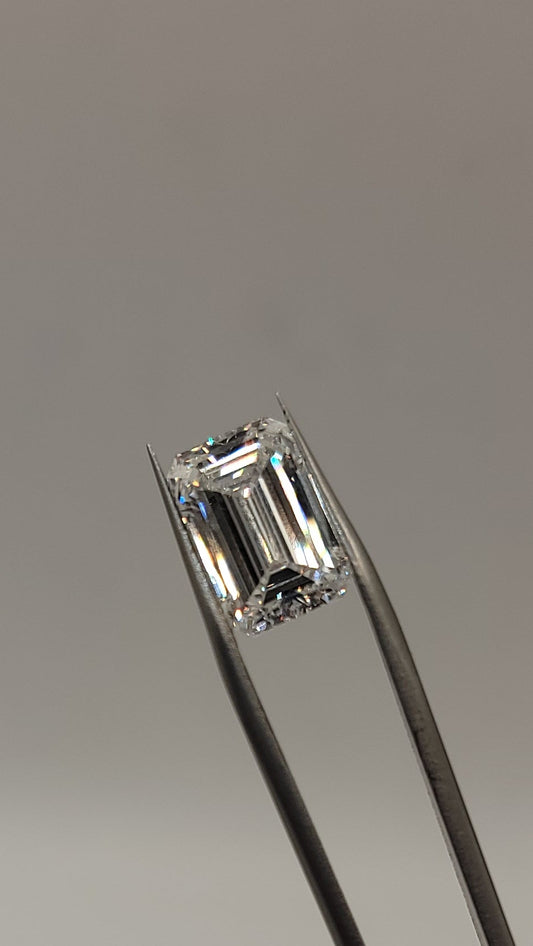 A single emerald-cut clear gemstone held in silver tweezers against a plain light gray background.
