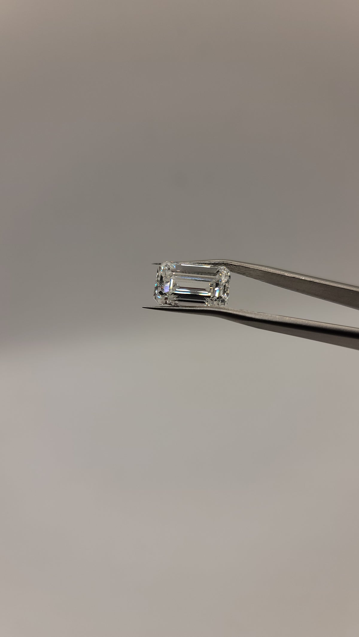 An emerald-cut clear gemstone held by silver tweezers against a plain light gray background.