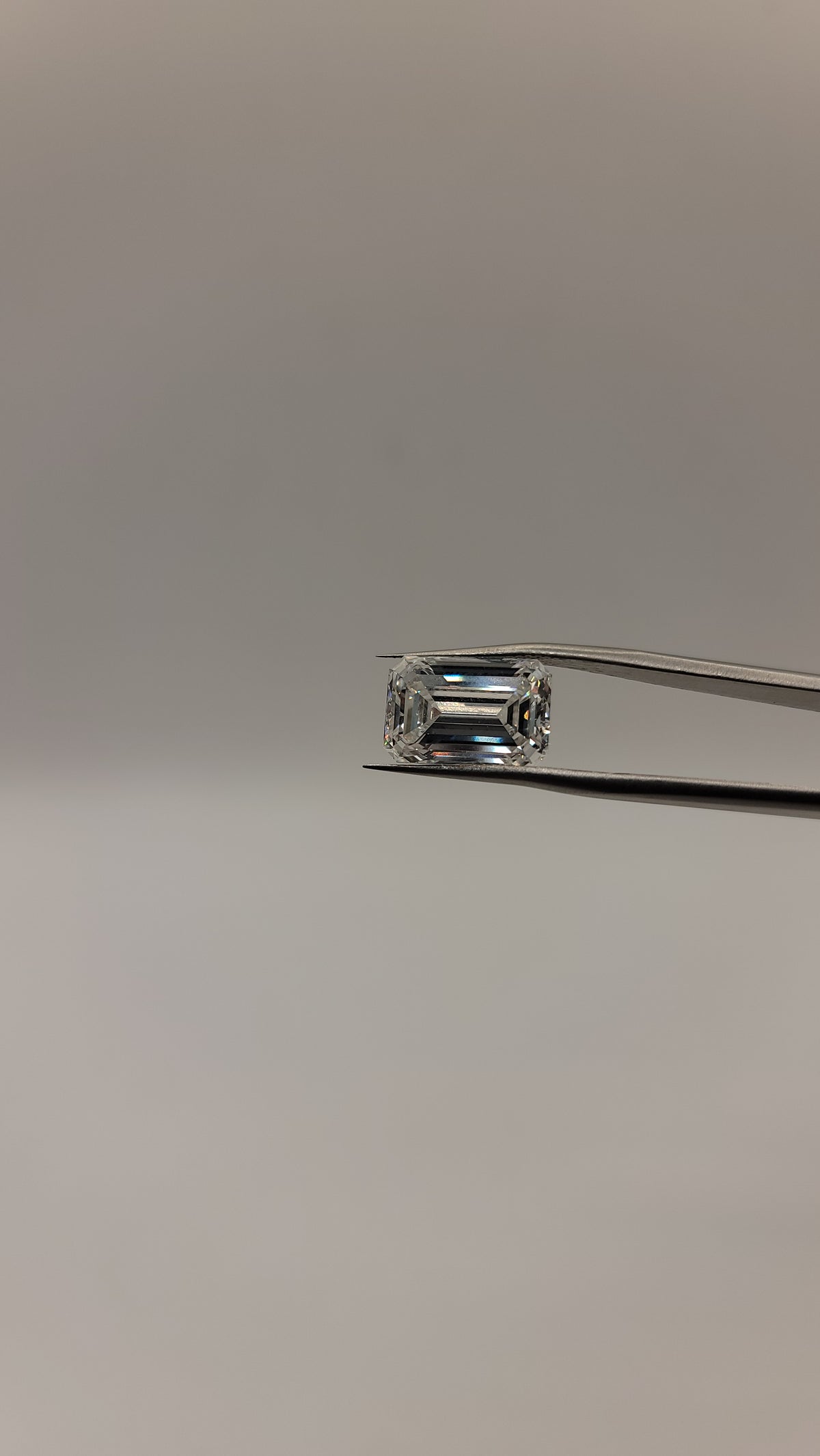 A rectangular emerald-cut clear gemstone held in a pair of metal tweezers against a plain light gray background.