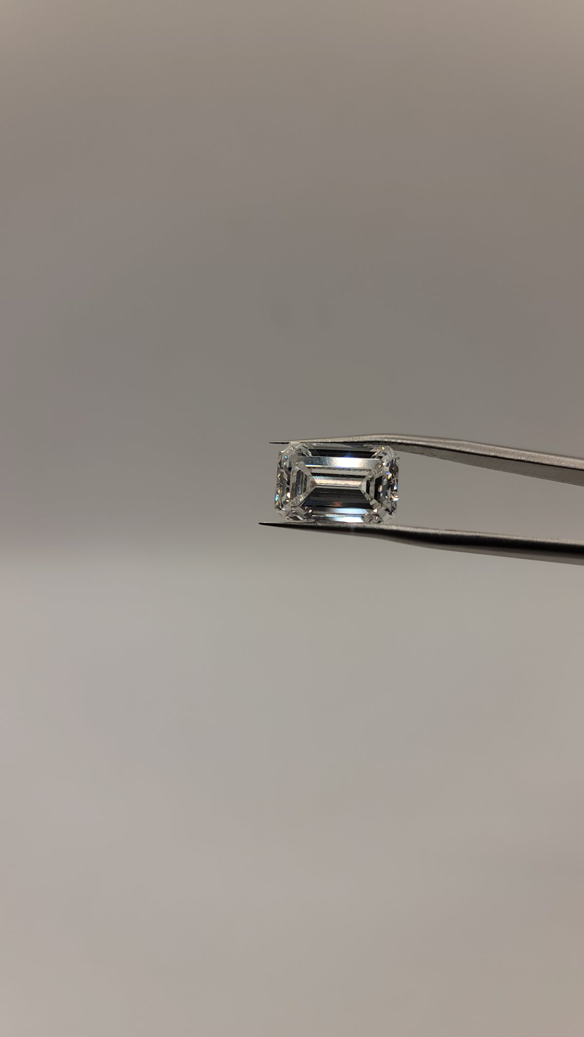 An emerald-cut clear gemstone held in silver-toned tweezers against a plain light gray background.