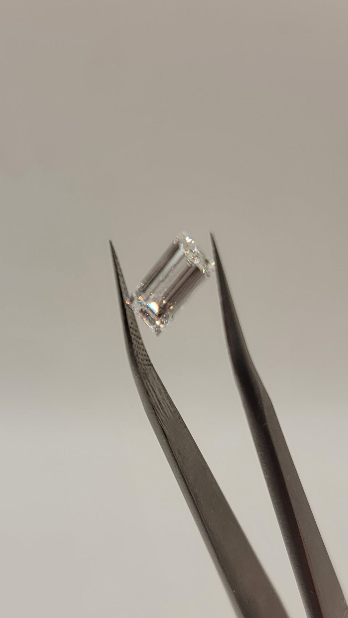 A rectangular clear gemstone held in a pair of metal tweezers, with a person's fingers visible at the bottom right against a plain light background.