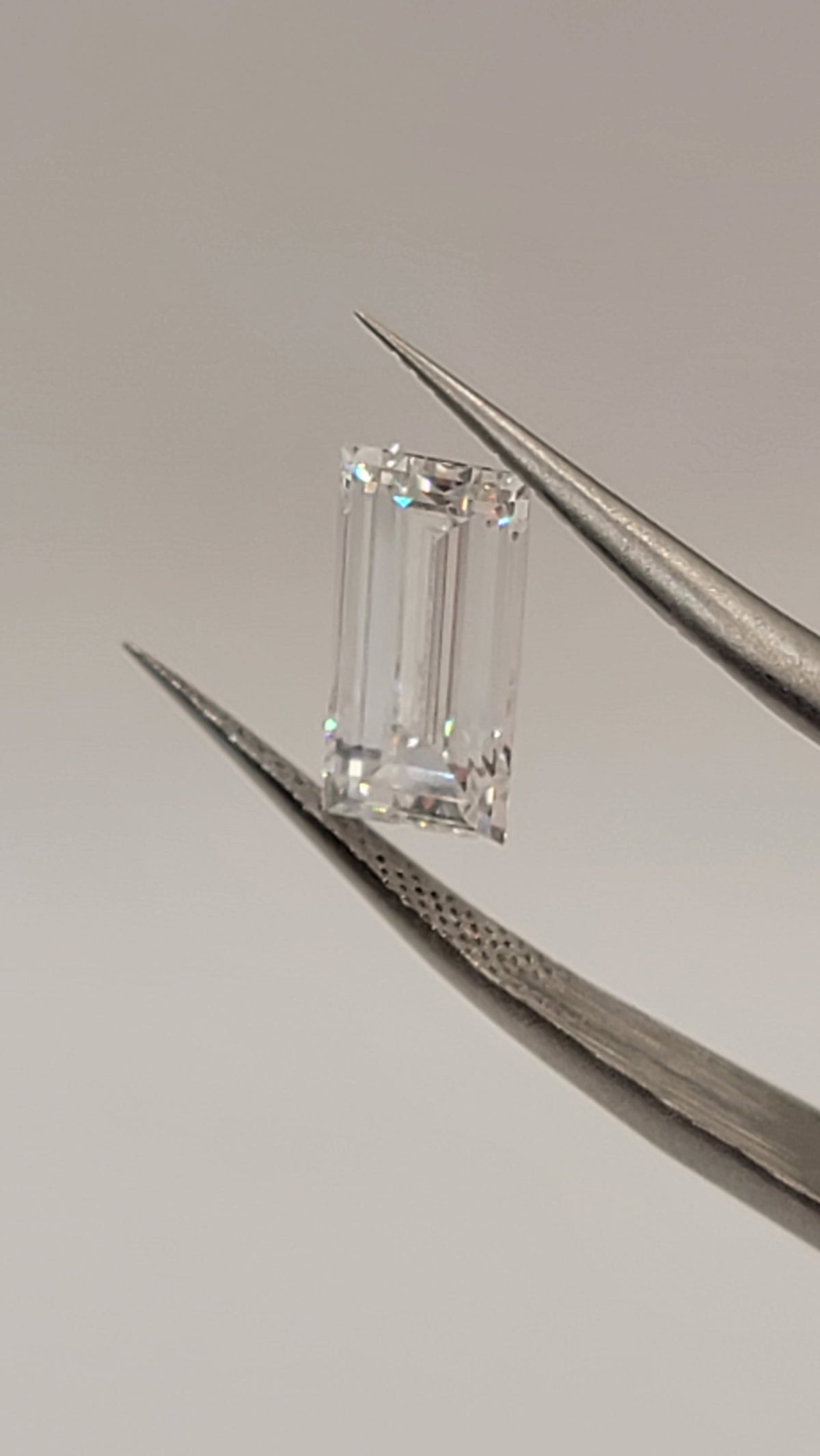 A rectangular emerald-cut clear gemstone held between two metal tweezers against a plain light gray background.