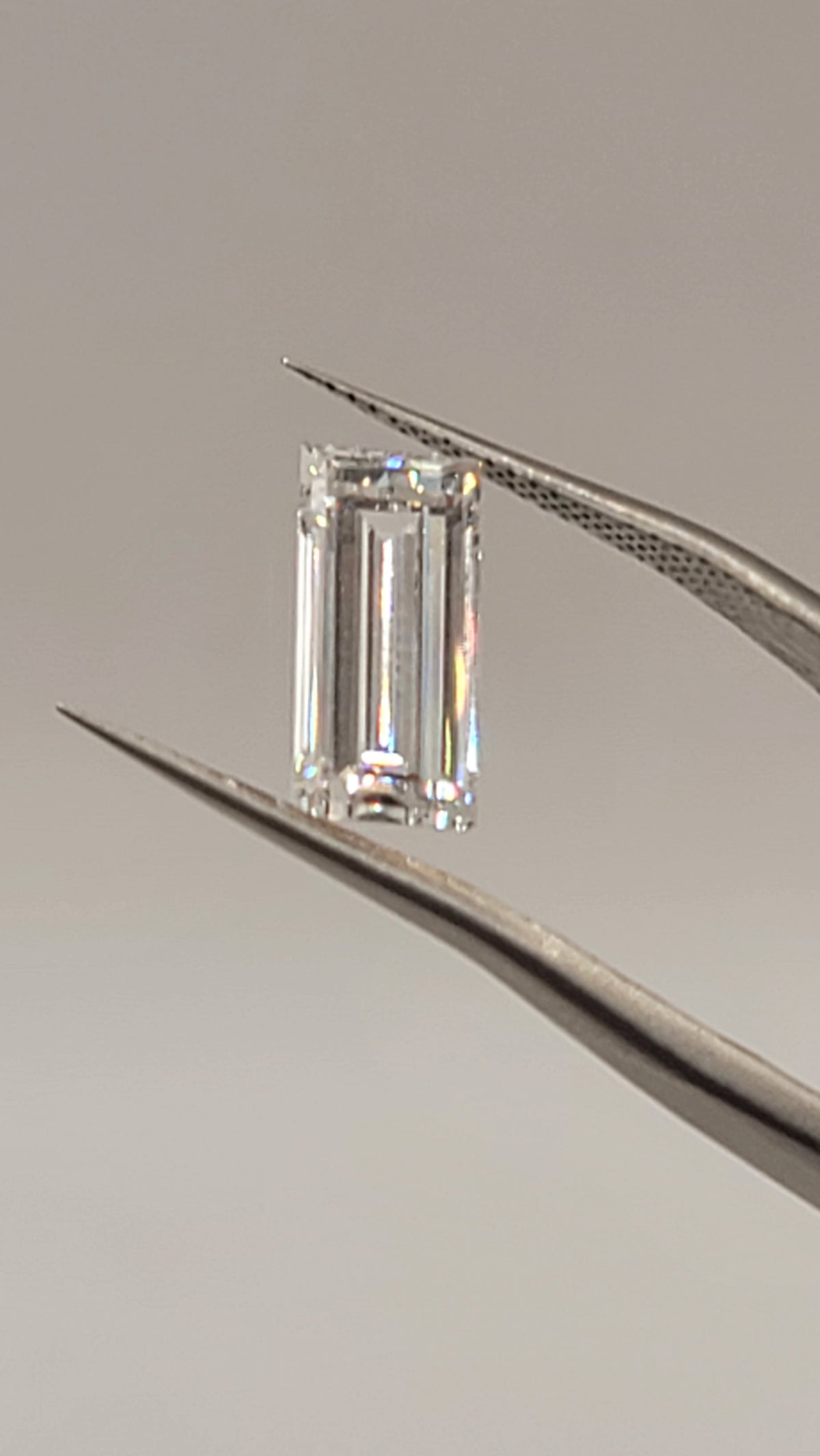 A rectangular emerald-cut clear gemstone held between metallic tweezers against a plain light gray background.