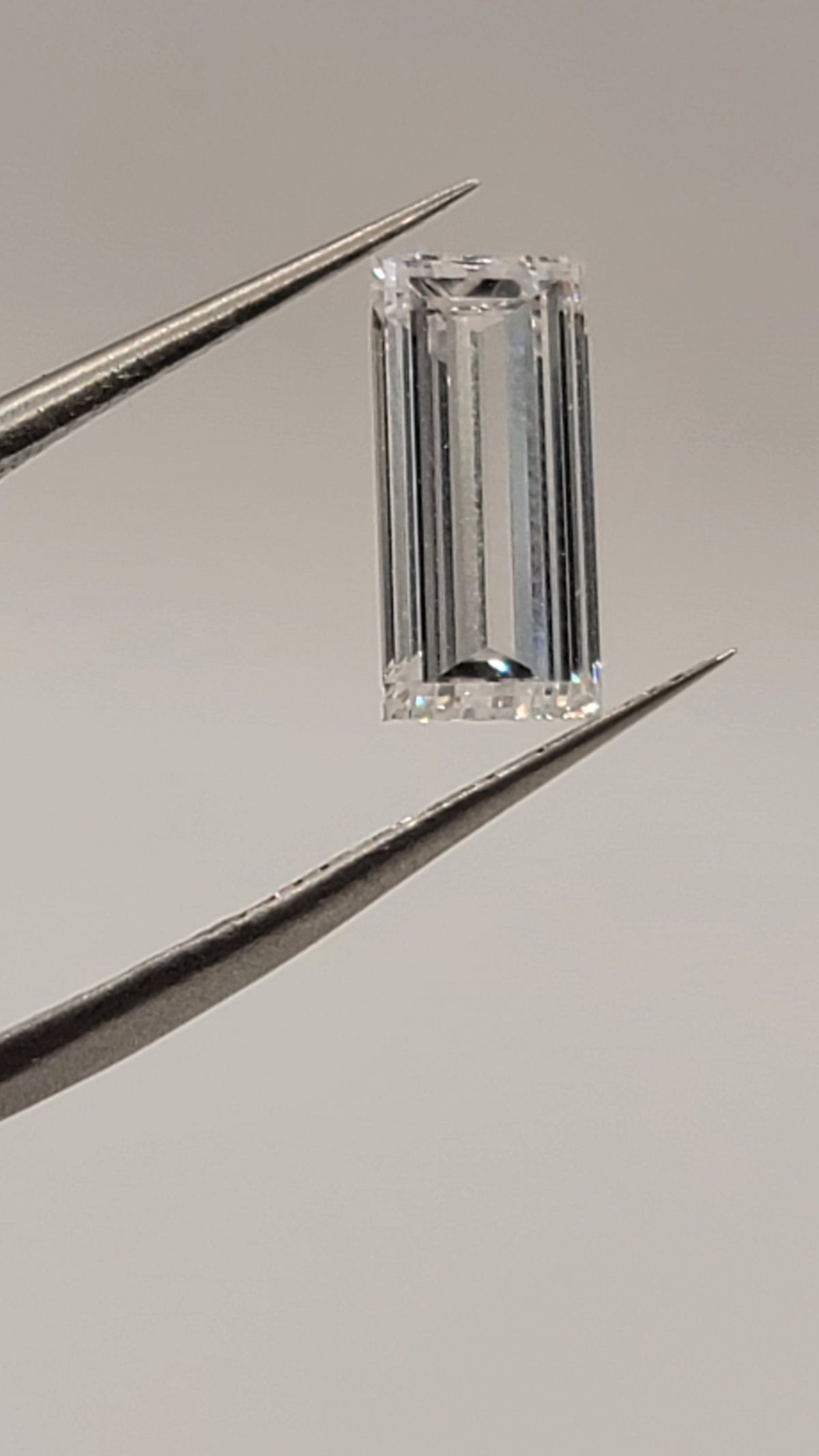 A rectangular emerald-cut clear gemstone held between metal tweezers against a plain light gray background.