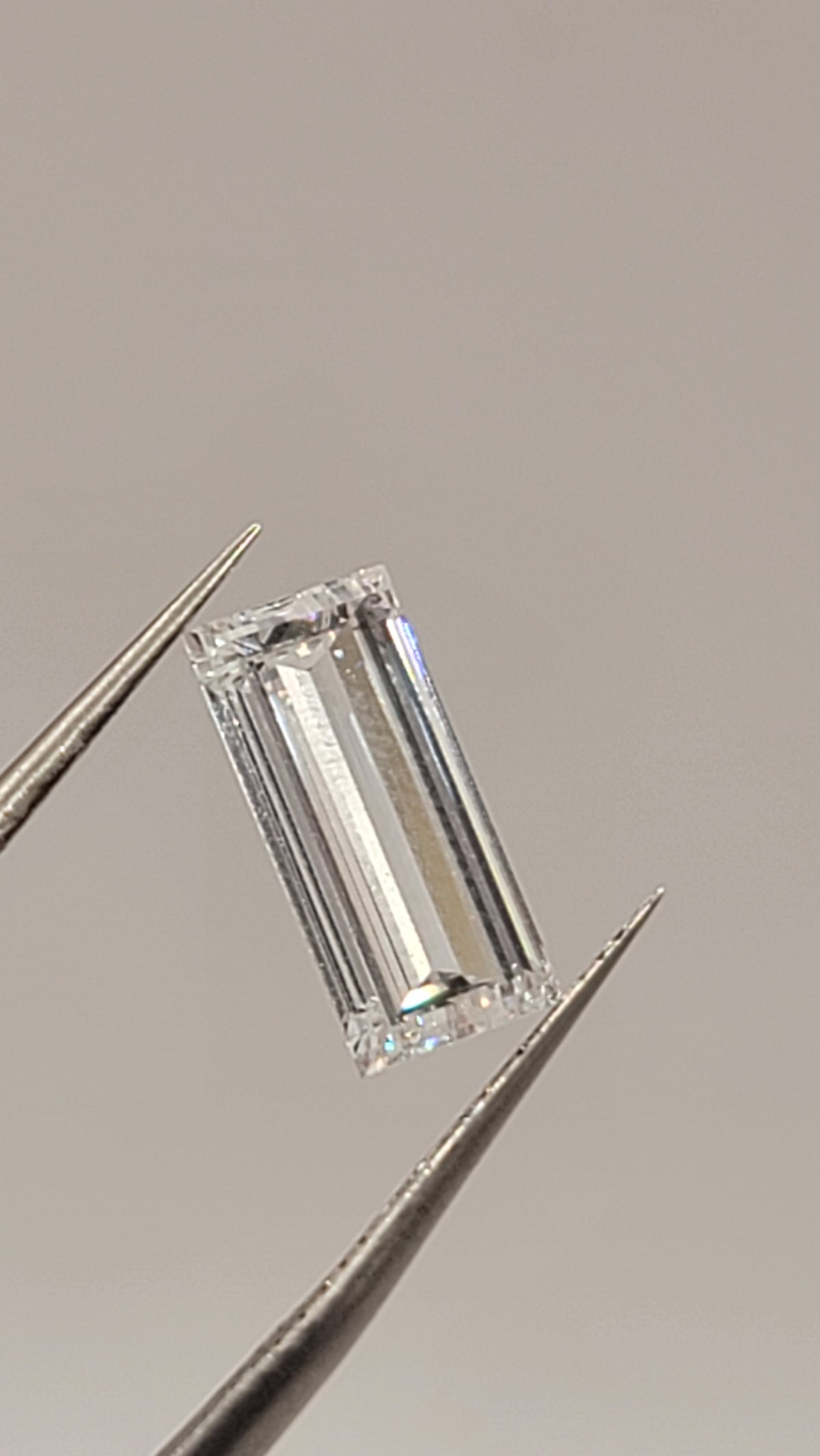 A rectangular emerald-cut clear gemstone held between metal tweezers against a plain warm gray background.