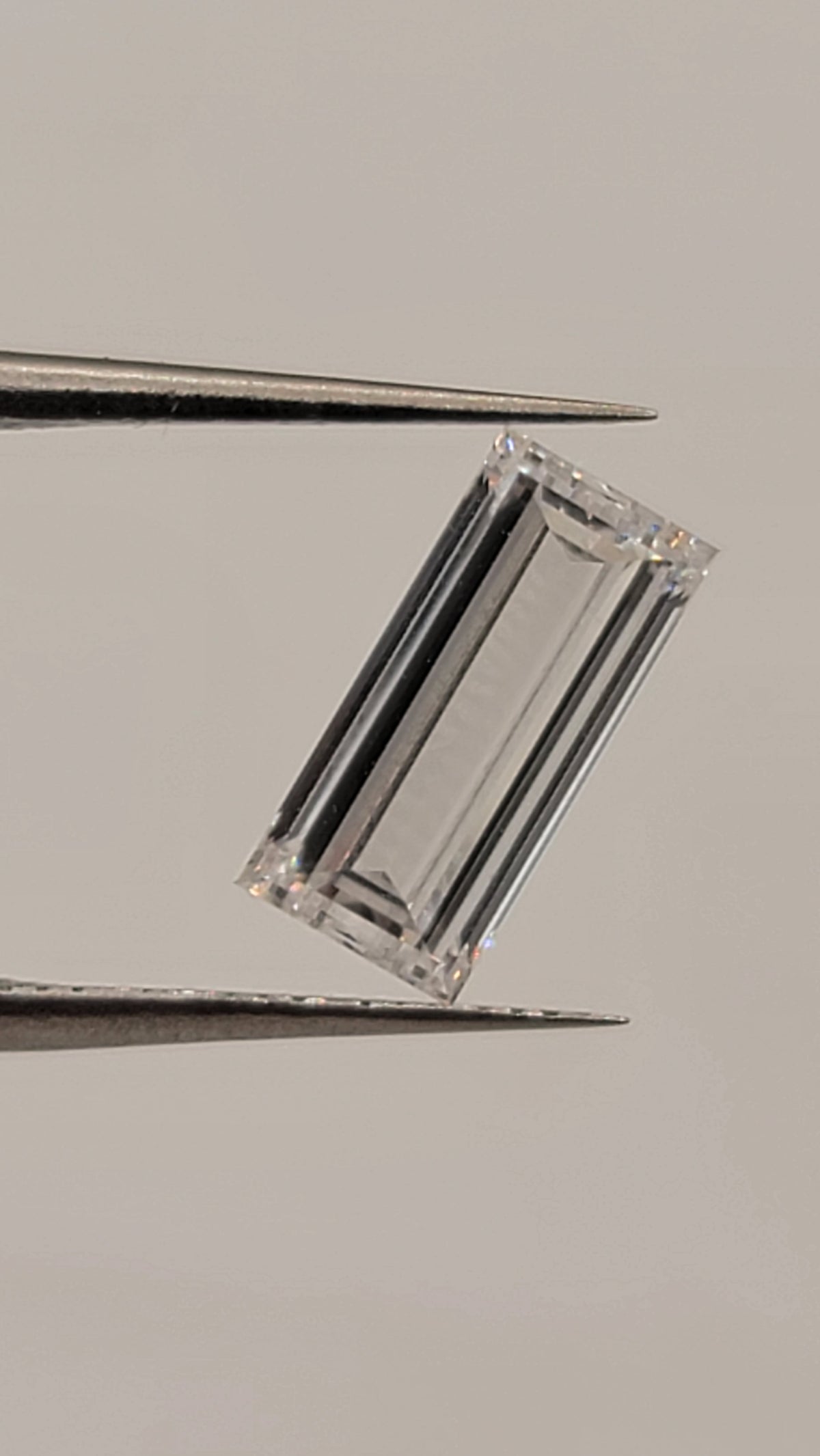 A rectangular clear gemstone held between metal tweezers against a plain light gray background.