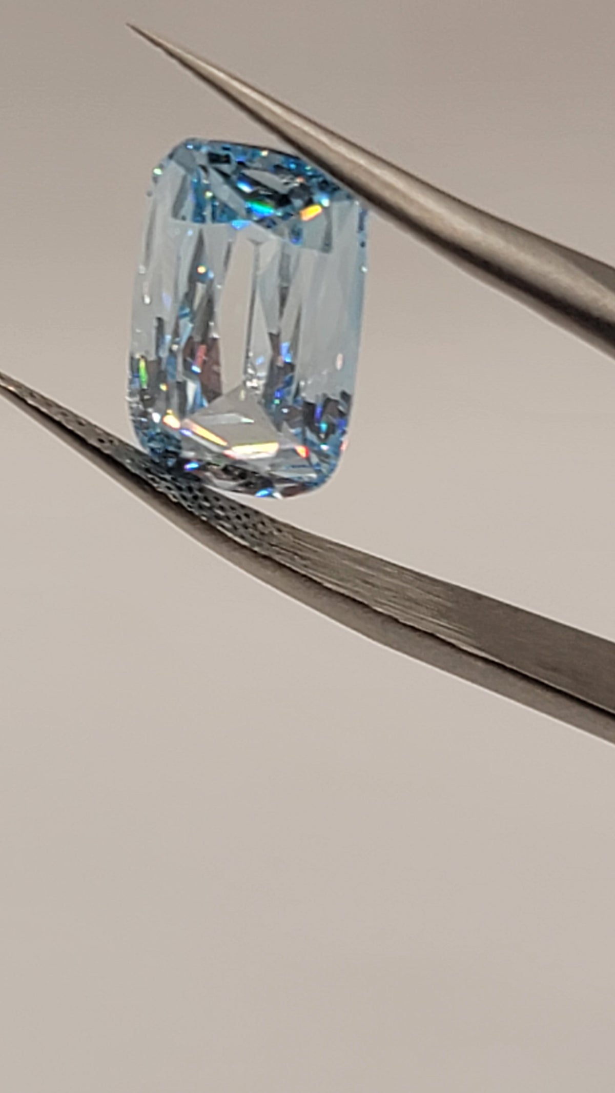 A light blue cushion-cut diamond held in silver-colored metal tweezers against a neutral light gray background.