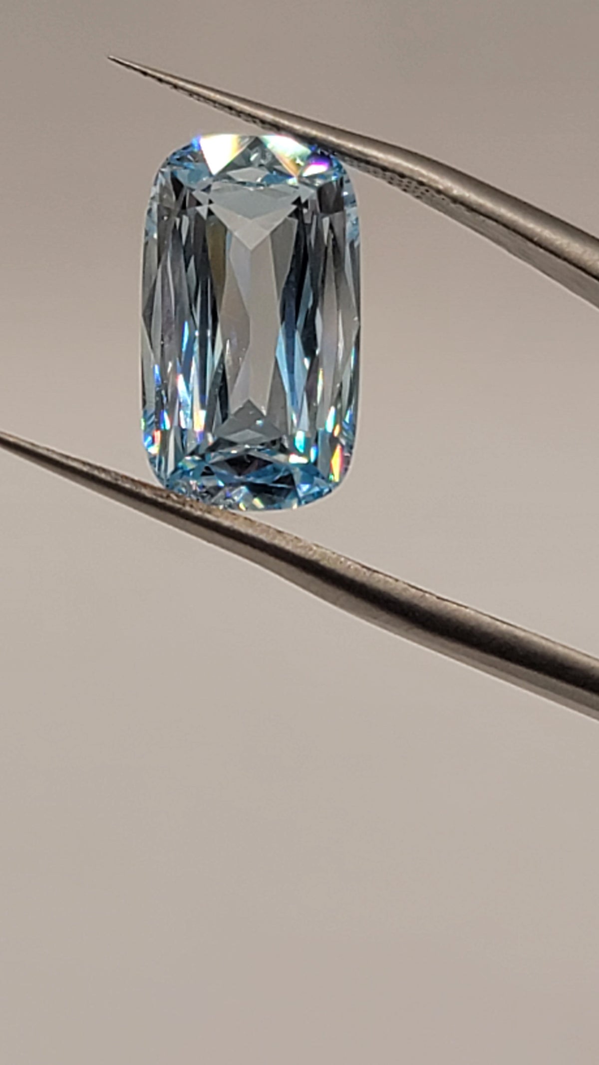 A rectangular cushion-cut light blue gemstone held between metal tweezers against a plain light gray background.