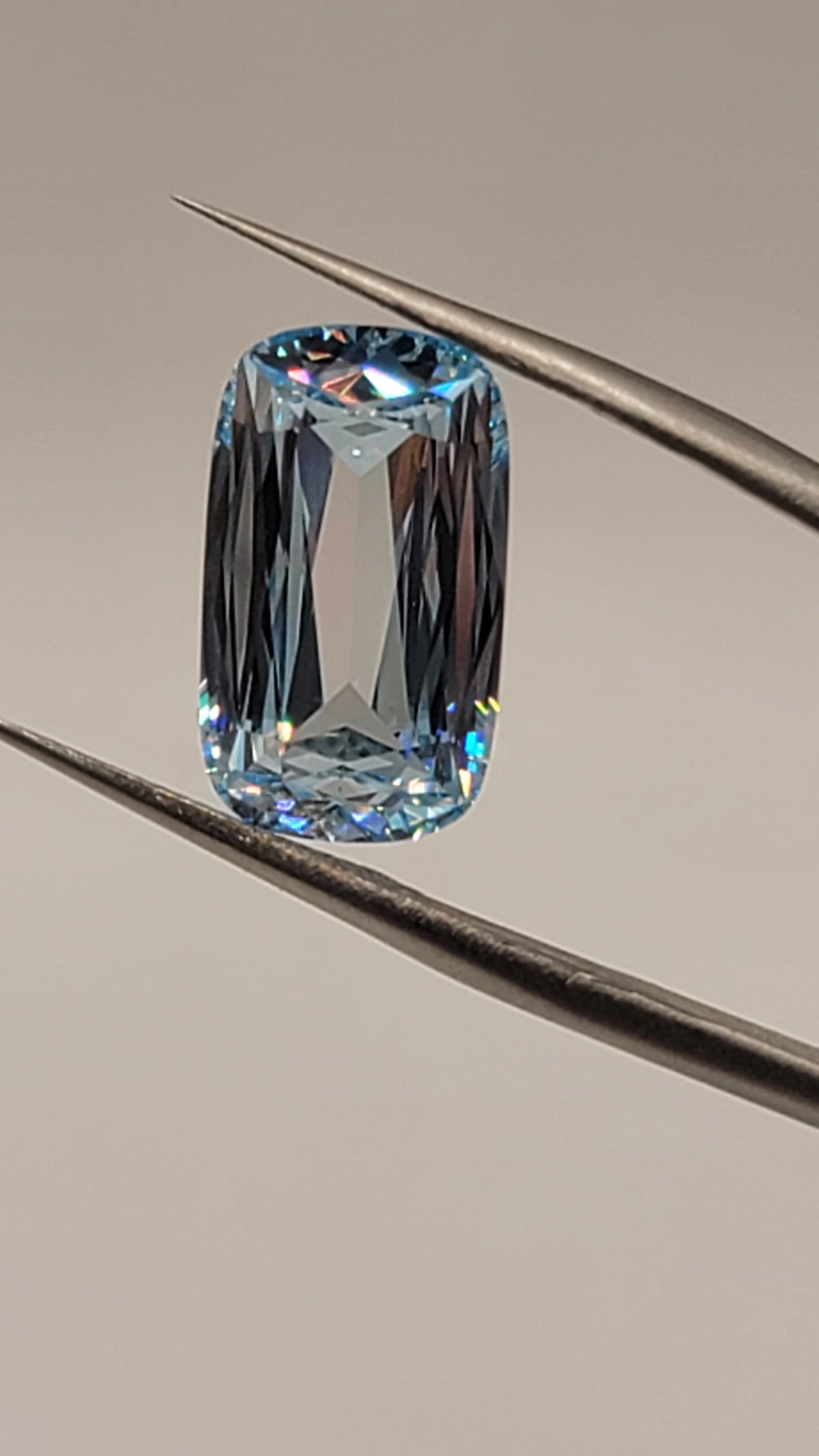 A loose cushion-cut light blue gemstone held between metal tweezers against a plain light gray background.