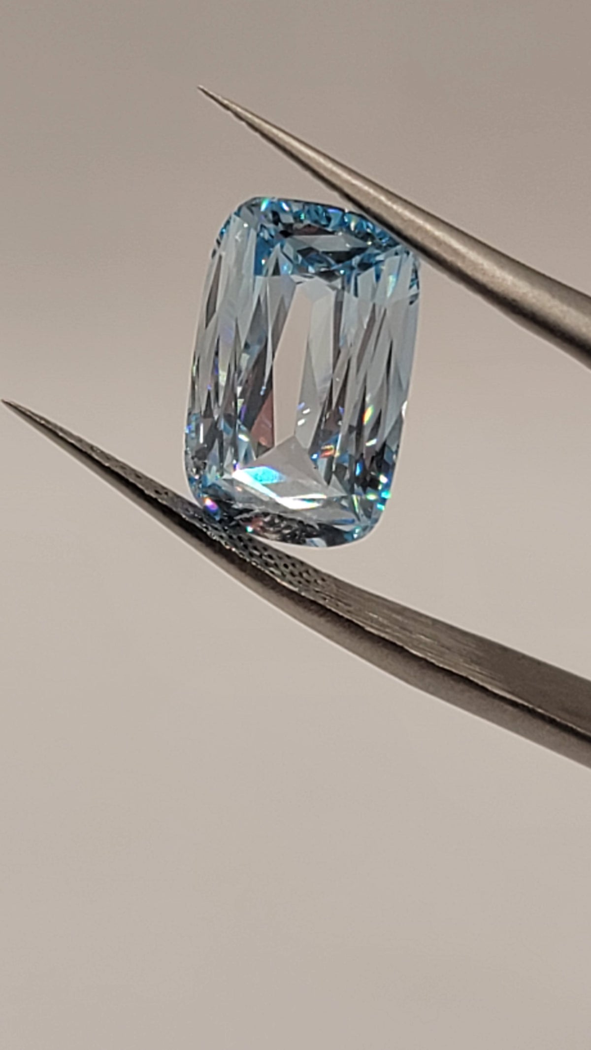 A rectangular cushion-cut light blue gemstone held in the jaws of metal tweezers against a plain light gray background.
