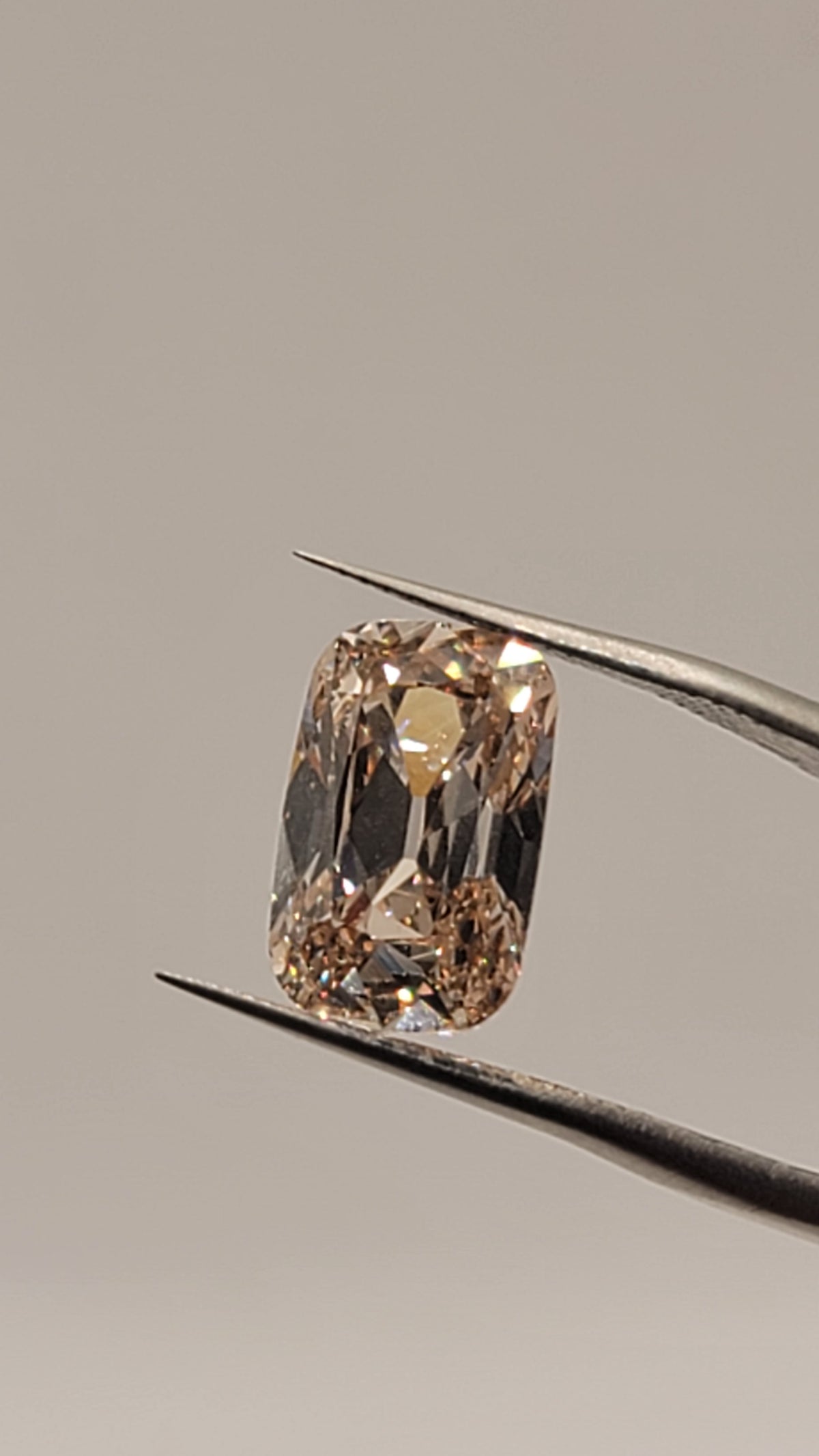 A cushion-cut diamond held in the jaws of metal tweezers against a neutral light gray background.