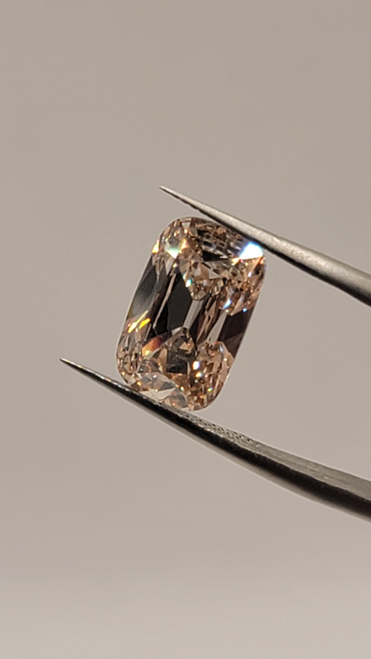 A cushion-cut diamond held in silver-colored metal tweezers against a solid neutral gray background.