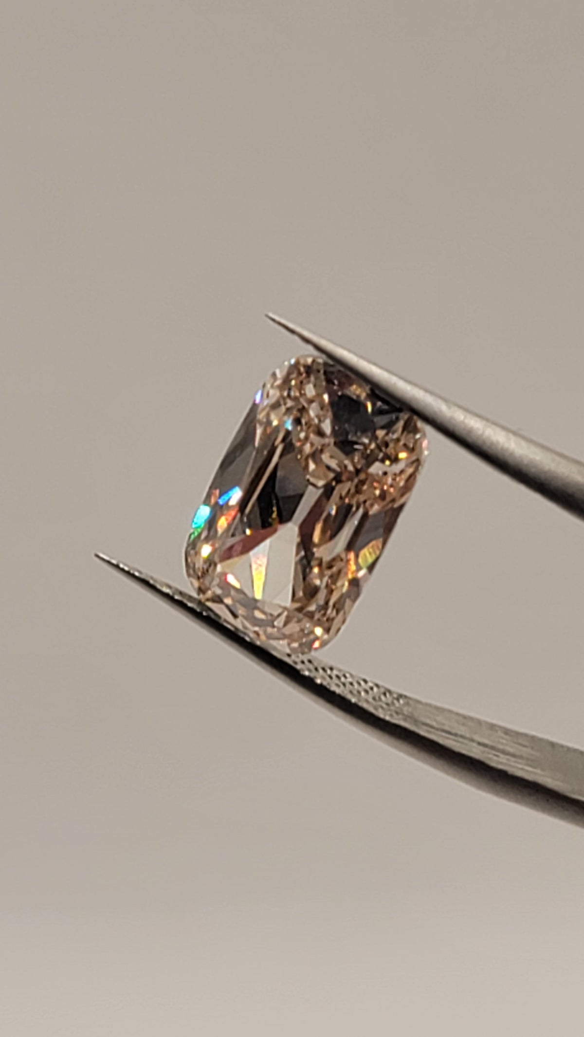 A rectangular cushion-cut diamond with a light pinkish-brown tint, held in the jaws of metal tweezers against a neutral gray background.
