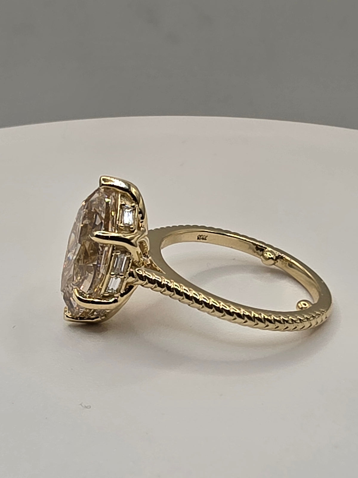 A yellow gold ring featuring a large oval-cut clear gemstone in a prong setting with baguette accents, displayed on a neutral light gray background.
