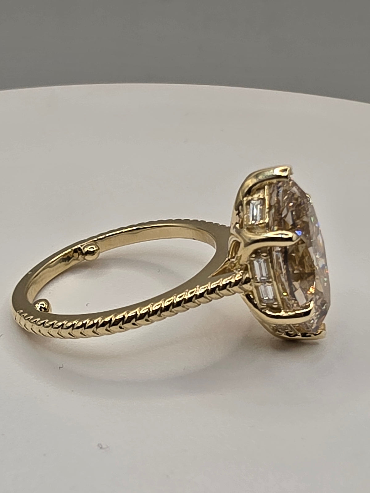 A yellow gold ring featuring a large oval-cut clear gemstone in a prong setting with smaller baguette-cut side stones, displayed on a neutral light gray surface.