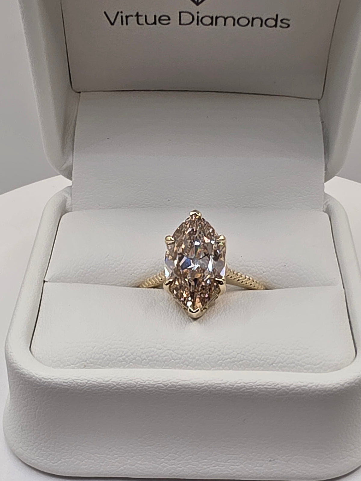 A yellow gold ring with a marquise-cut clear diamond set in a white velvet jewelry box branded 'Virtue Diamonds'.