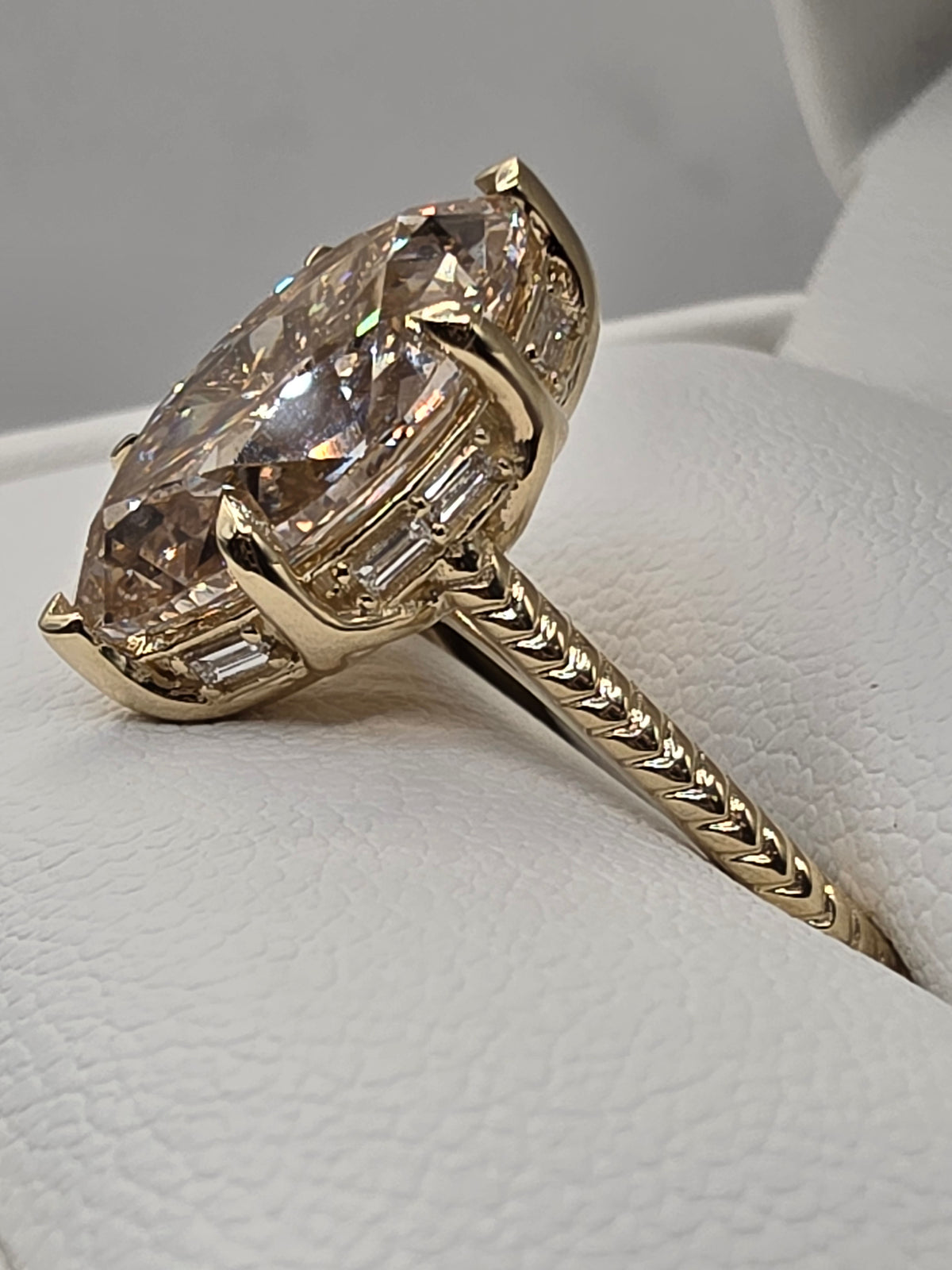 A yellow gold ring featuring a large oval champagne-colored center stone with baguette side stones, displayed on a textured white ring stand.