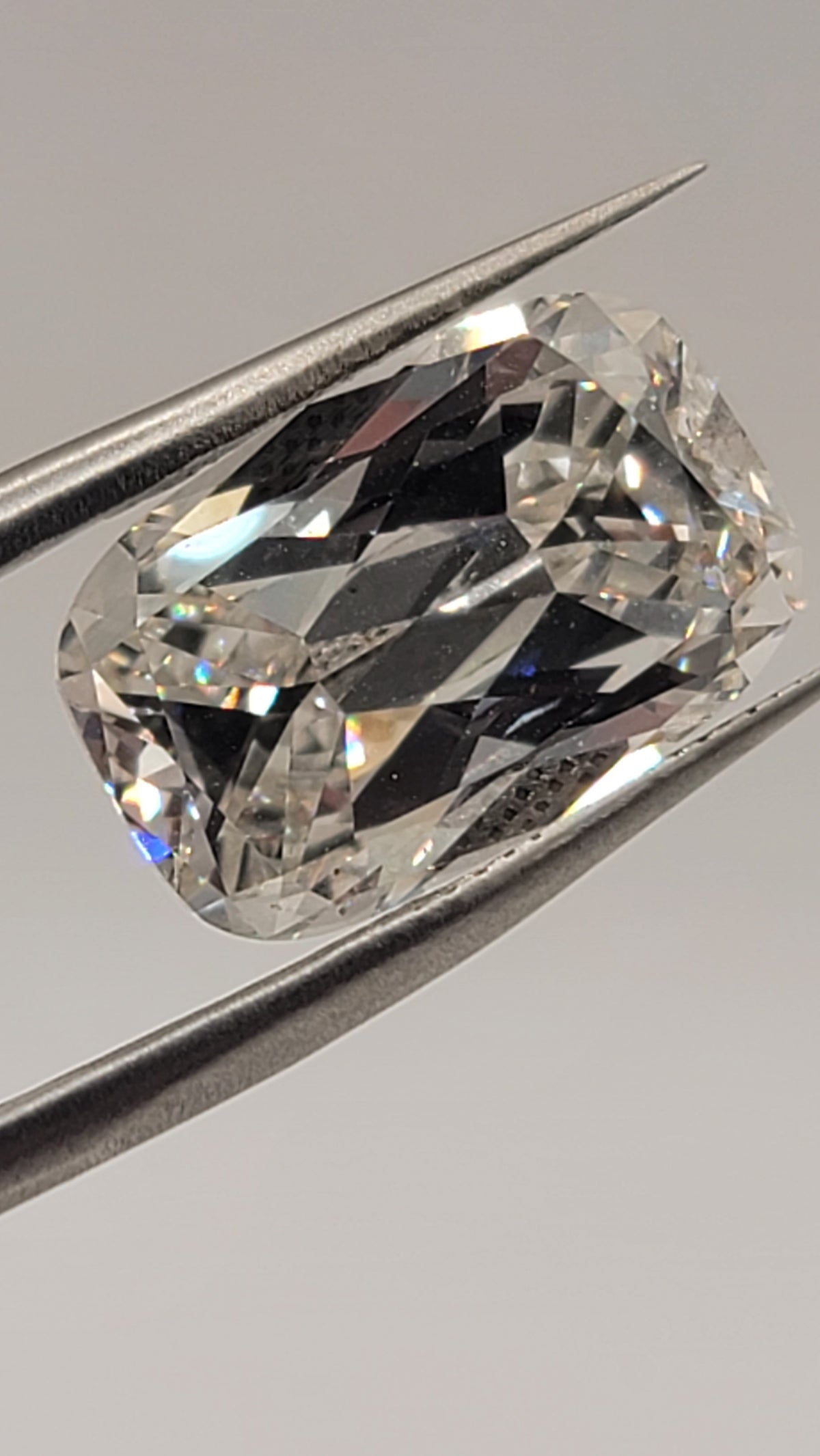A loose cushion-cut clear gemstone held between metal tweezers against a light gray background.