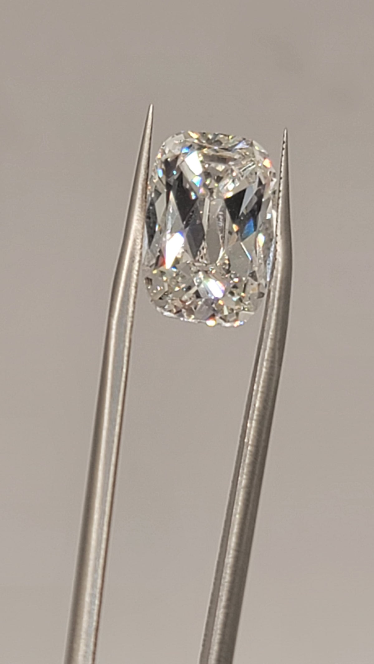 A loose cushion-cut diamond held in the tips of metal tweezers against a plain light gray background.