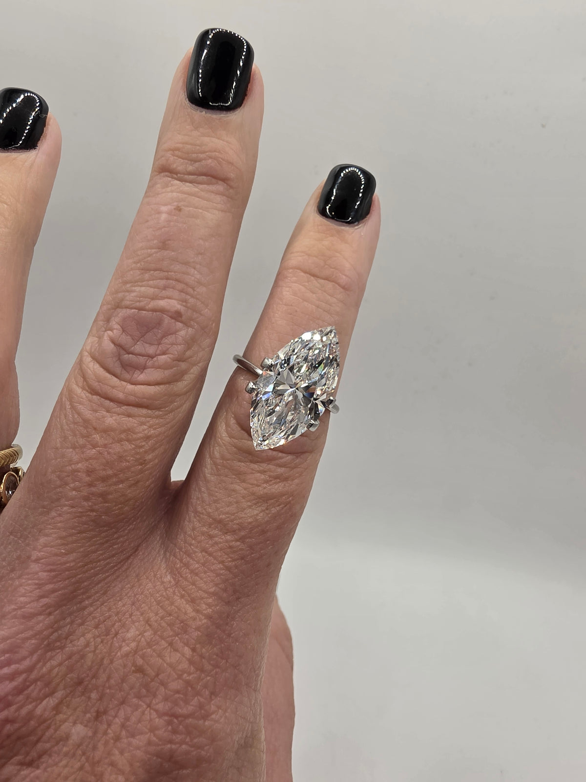 A close-up of a hand wearing a silver ring with a large marquise-cut clear gemstone. The nails are painted black, and the background is a plain light gray.